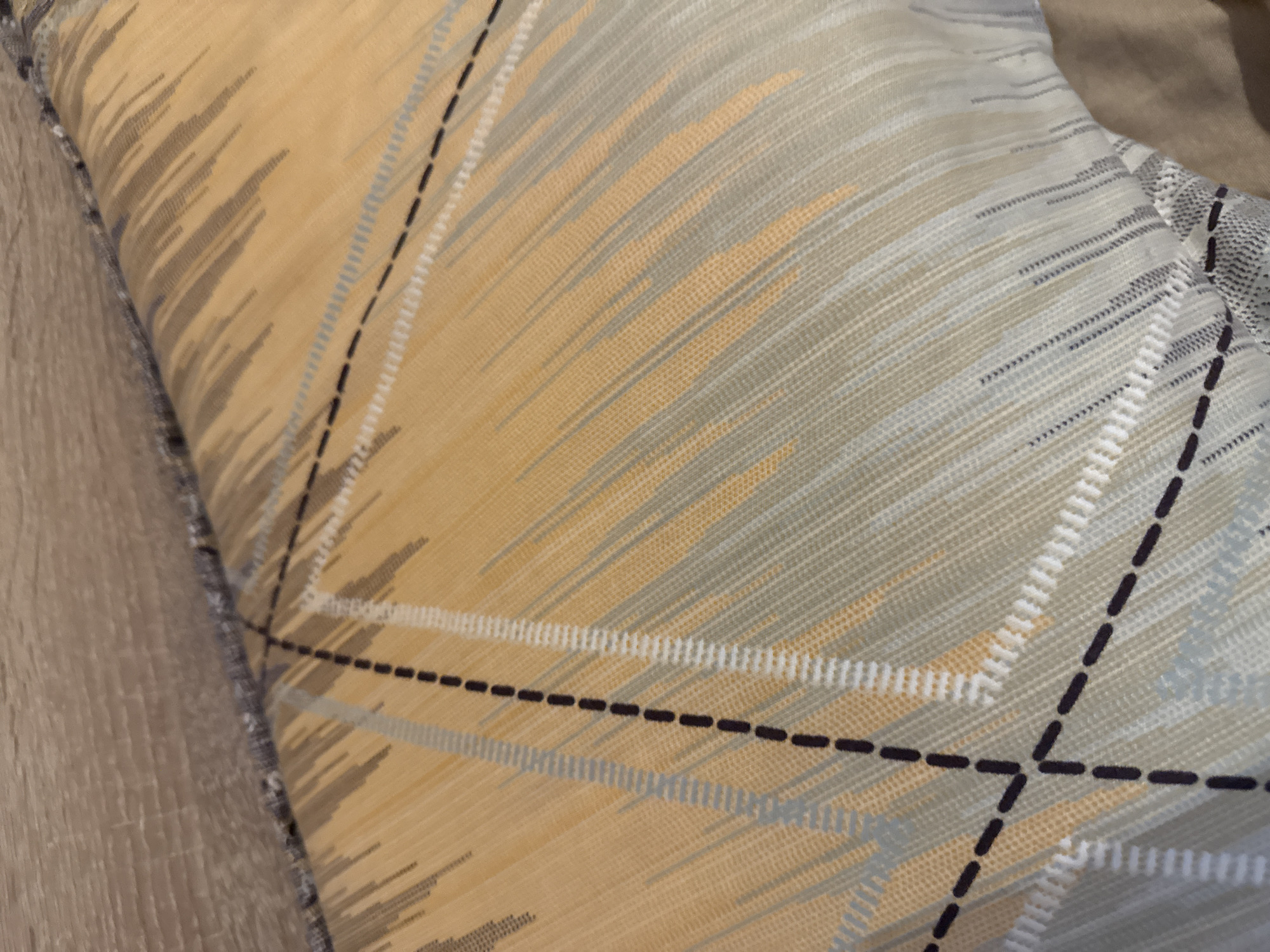 A close-up view reveals a textured pillow with a distinctive pattern. The pillow's fabric features a geometric design consisting of intersecting lines, some rendered in a dashed, dark brown hue. The spaces between these lines are filled with alternating bands of light gray and a warmer, beige color, contributing to a complex and visually rich surface. The pillow appears slightly creased and folded, suggesting it is in use or has been recently handled. The image does not provide information about the location in Lovech, Bulgaria.