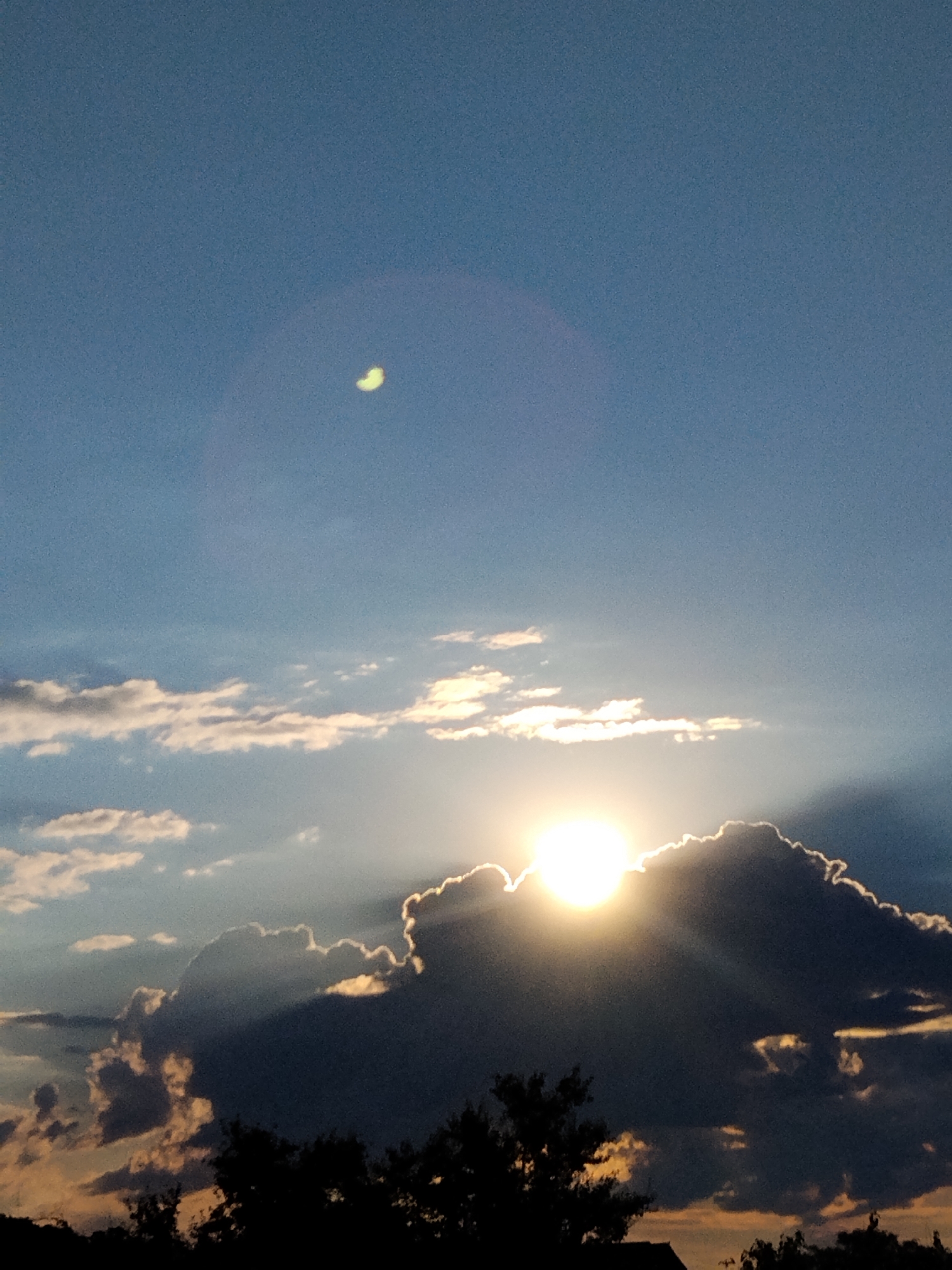 The image captures a dramatic sunset over Ustyvytsia, Ukraine. The sun, a brilliant orb, peeks from behind a dark, imposing cloud, casting golden rays that dramatically illuminate the edges of the clouds and create visible light beams. In the upper portion of the sky, a crescent moon is faintly visible against the clear blue expanse. Silhouetted trees and the dark outlines of rooftops are visible at the bottom of the frame, suggesting a residential area. The overall atmosphere is one of a powerful and dynamic natural event.