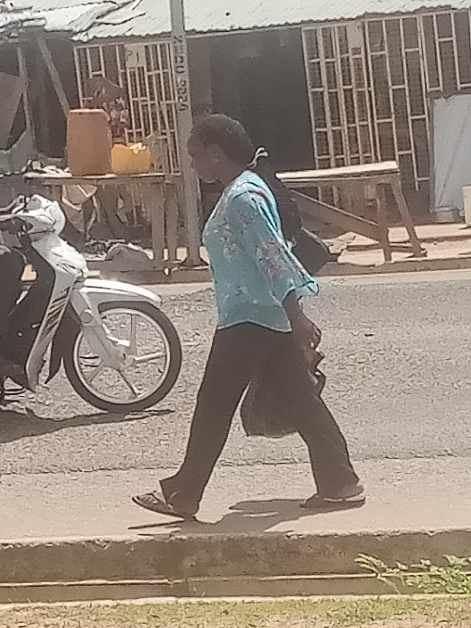 A person is walking on a sidewalk in what appears to be a street scene in Jalingo, Nigeria. The person is wearing a light blue, patterned long-sleeved shirt and dark pants, with sandals on their feet. They are carrying a dark bag over their shoulder. To the left of the person is a white motorcycle, partially visible. In the background, there is a building with a corrugated metal roof and a metal grate structure. There are also some tables and containers, including a large orange one, suggesting a market or roadside stall. The lighting suggests it is daytime, likely sunny, as evidenced by the shadows. The overall scene depicts a person going about their day in an urban or semi-urban environment. No discernible emotions are evident from the image. There is some faint text on a pole in the background, but it is illegible.