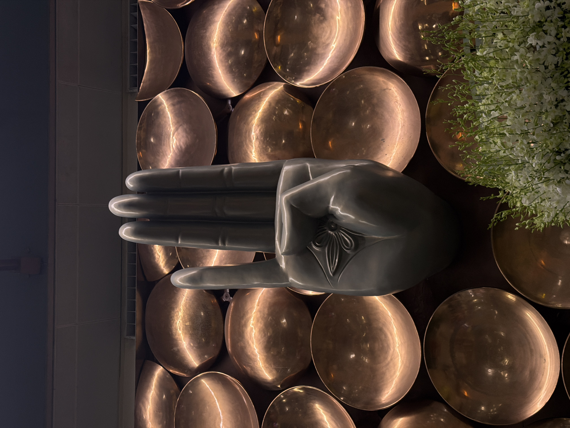 A close-up, low-angle shot captures a large, grey, stylized sculpture of a hand with its index and middle fingers raised. The hand sculpture is positioned in front of a wall adorned with numerous round, copper-colored decorative bowls. The bowls are arranged in a honeycomb pattern and reflect warm light, creating a textured background. In the lower right corner of the image, a lush arrangement of delicate white flowers with green stems is visible, partially obscuring some of the bowls. The lighting is dramatic, with strong highlights on the hand sculpture and the copper bowls, and a dark, undefined background above the bowls. The scene appears to be indoors, possibly an art installation or decorative element in a public space or private residence in Delhi, India.