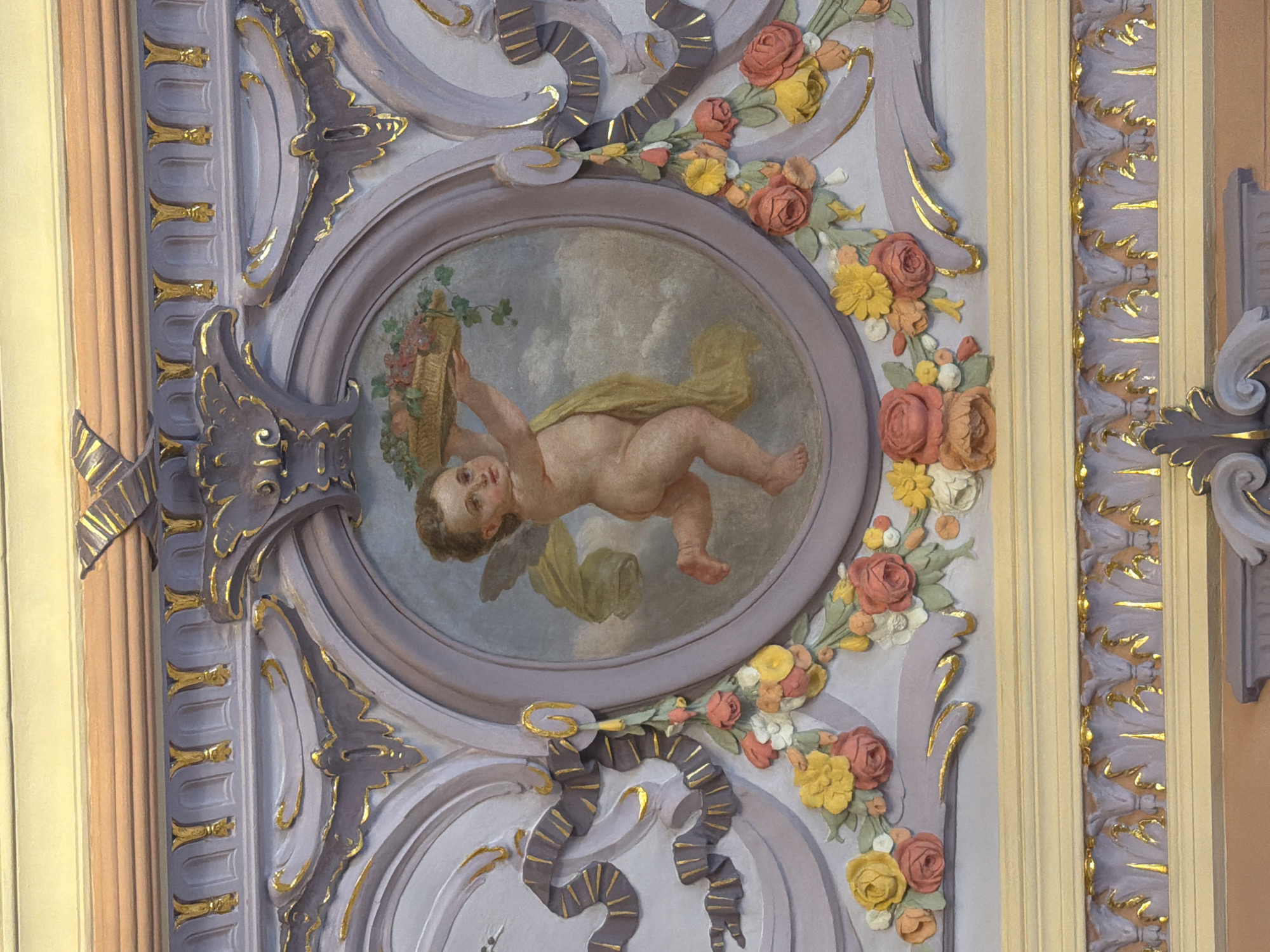 This is a photograph of a decorative ceiling panel featuring a painted cherub and ornate stucco work. The cherub, depicted as a chubby infant with small wings, is reaching upwards, holding a woven basket overflowing with grapes and other fruits. The cherub is draped in a yellow cloth that flows behind it. The painting is framed by a circular stucco relief that is part of a larger, elaborate decorative architectural element. This stucco work is predominantly light purple and adorned with gold accents, featuring swirling patterns, floral motifs like roses and smaller blossoms, and ribbon-like elements. The surrounding architecture is also decorated with similar stucco details and gilded trim. The context of this image is likely an interior architectural space, possibly a theater, palace, or grand hall. Specific location information for Nizhny Novgorod, Russia, is not discernable from the image itself.