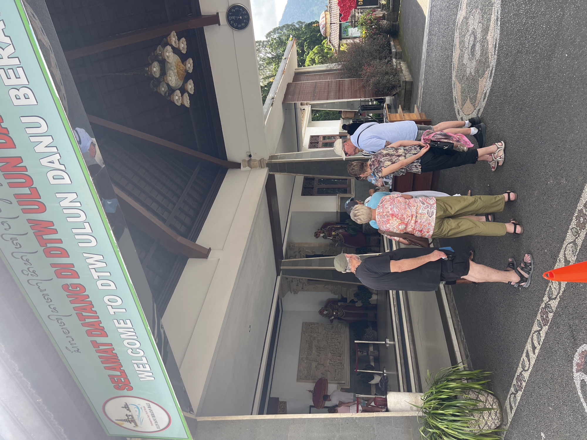 The image shows a group of tourists standing in front of the entrance to DTW Ulun Danu Beratan in Baturiti, Indonesia. There are four visible tourists lined up, wearing casual clothing, including sandals and hats. The entrance features a welcome sign in both Balinese and English, indicating the name of the attraction. The building has a traditional Balinese architectural style, with a tiled roof, wooden beams, and decorative carvings. A clock hangs on the building's exterior, and there are potted plants and decorative stones along the walkway. The ground is asphalt with decorative inlaid patterns. An orange cone is visible to the right of the image.