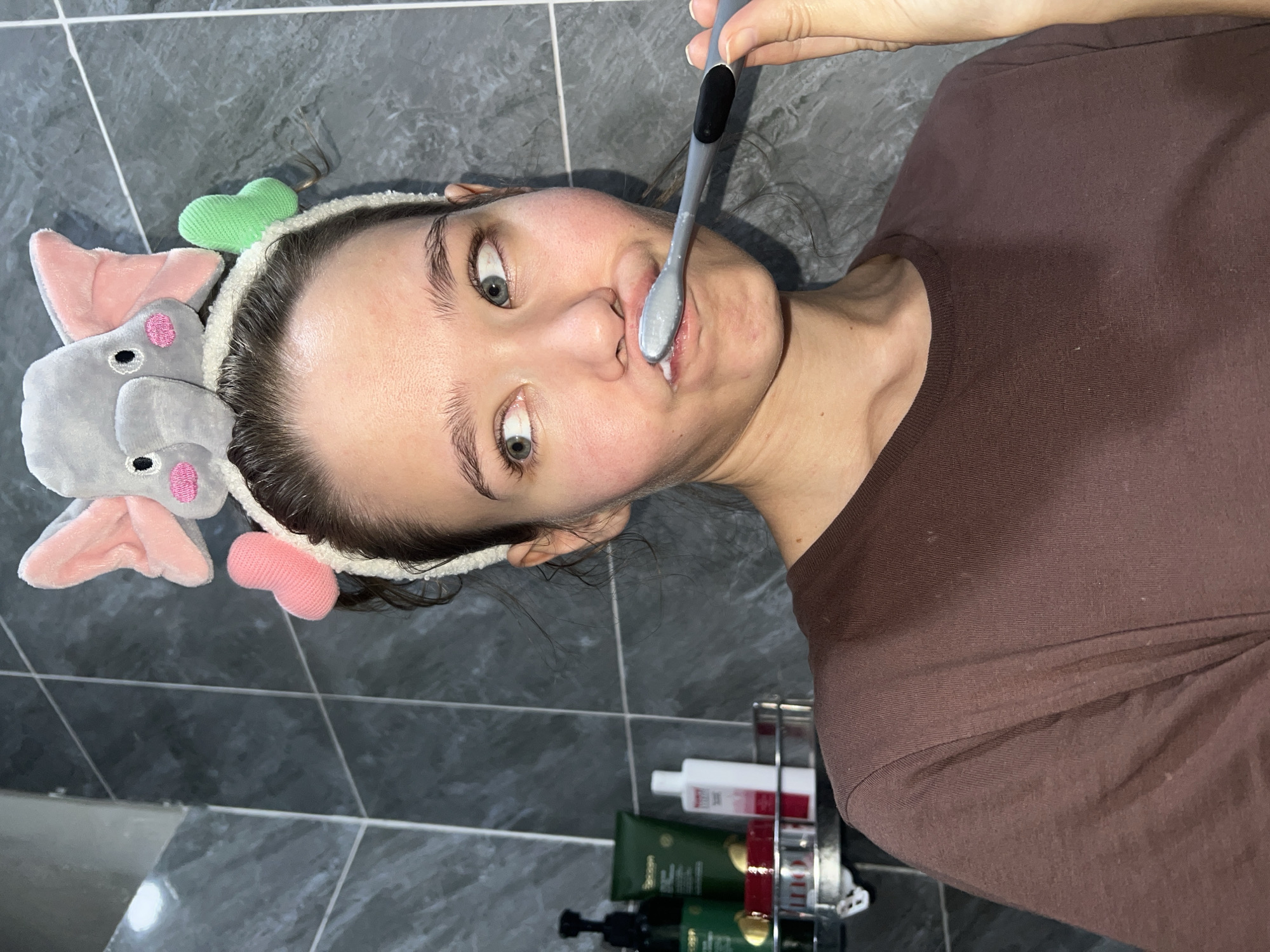 A young woman is depicted in a close-up, eye-level shot, brushing her teeth in a bathroom in Nam Nha Trang Ward, Vietnam. She has wide, open eyes that look directly at the viewer, conveying a surprised or playful expression. Her mouth is slightly open, revealing a toothbrush with white foam or toothpaste.
She is wearing a plain brown short-sleeved t-shirt. A white, fluffy headband holds her dark hair back from her face. The headband is adorned with novelty elements: soft grey elephant ears and trunk on the left side, a light green knitted heart on top, and a pink knitted heart on the right side.
The background features dark grey or black square tiles with white grout lines, consistent with a bathroom setting. To her right, a metallic shower caddy holds various toiletries. Visible items include two dark green bottles, one clearly labeled "BIOLAGE" and "MATRIX," a white bottle with a red label, and a smaller red bottle. The lighting is soft and ambient.