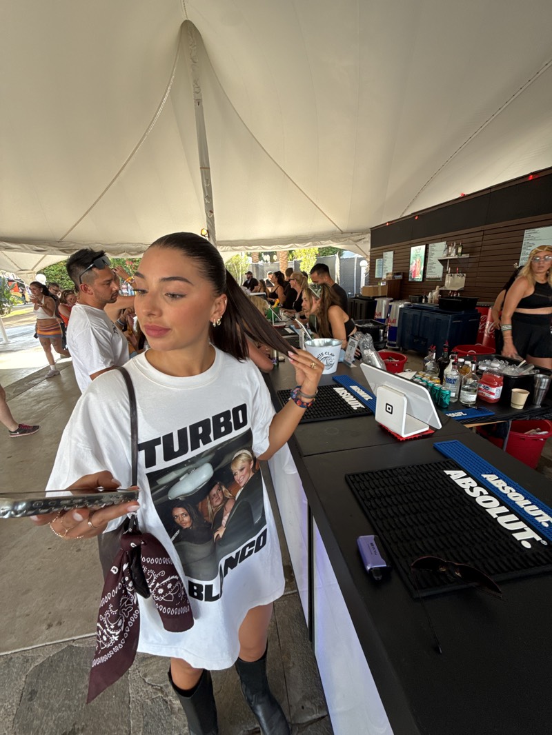 The image shows a young woman standing at a bar inside a tent-like structure in Indio, United States. She has her hair in a ponytail and is wearing a white t-shirt with "TURBO BLANCO" printed on it, along with a photo of several young women in a car. She is also wearing black boots, and a shoulder bag with a dark bandana tied to it. She is holding a cell phone in one hand and touching her ponytail with the other.
Behind her is a black bar counter with "ABSOLUT" branded mats and various bottles and bar equipment. Other people are standing around the bar, some waiting to be served. The environment suggests an outdoor event, possibly a festival or fair, held in Indio, California.