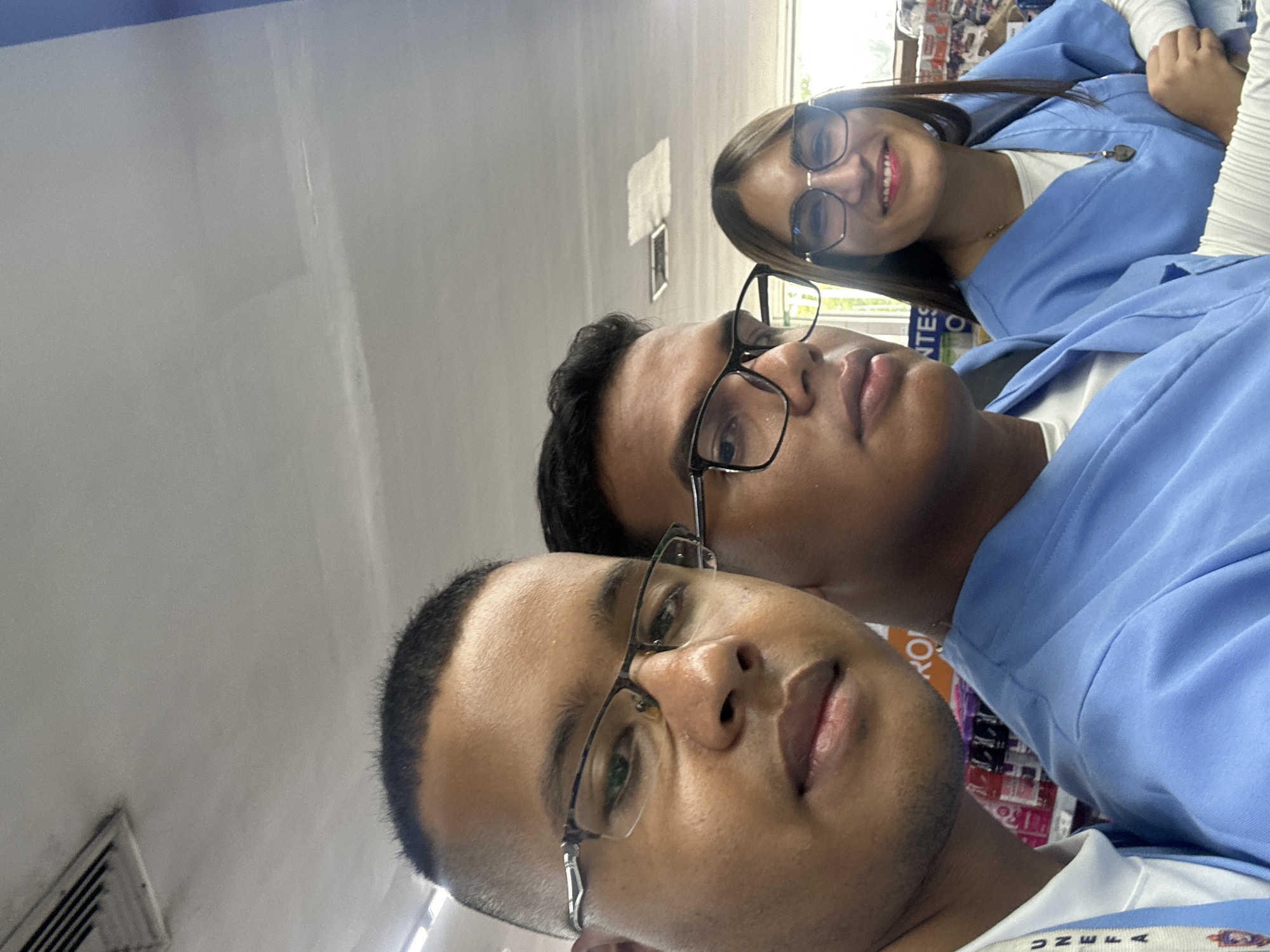 Three people are taking a selfie. Two males with short dark hair and glasses are in the foreground, with the woman smiling in the background. All three are wearing light blue shirts, possibly uniforms. The background is a blurred retail or office space. The location is likely Parroquia Ocumare, Venezuela, based on a label visible on one of the shirts.