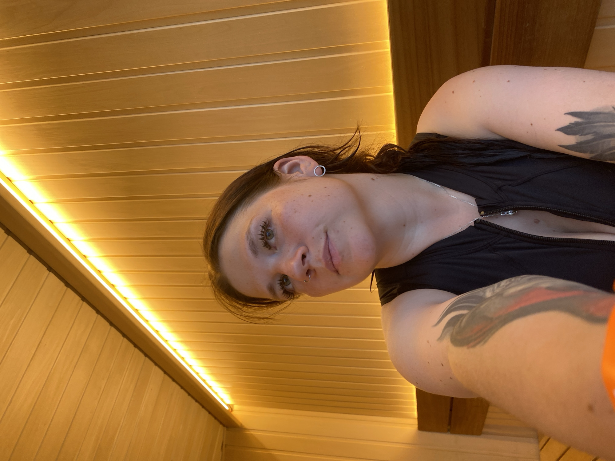 A young woman with dark hair and light eyes poses for a selfie in a wooden sauna. She is wearing a black zippered vest and a silver necklace. She has tattoos on both arms. The sauna has wooden walls and ceiling, with a strip of yellow LED lighting along the top of the wall. A wooden bench is visible in the foreground. The location is Votkinsk, Russia.