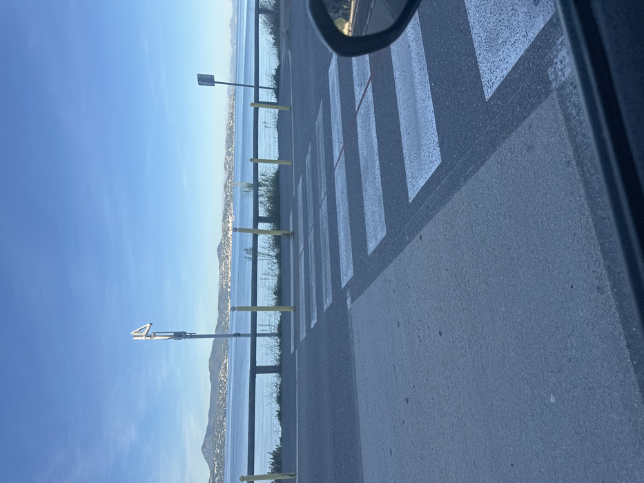 The image depicts a coastal road scene in Gassin, France, viewed from inside a vehicle, with a portion of a side mirror visible on the right. In the foreground, a dark asphalt road features a pedestrian crosswalk marked by white rectangular stripes. A lighter grey pavement or curb area is adjacent to the road on the left. The midground presents a black guardrail running horizontally along the road's edge, punctuated by several yellow vertical bollards. Sparse green vegetation grows beneath the guardrail, bordering a body of calm blue water. Two metallic poles stand along the roadside: a taller pole with an angular sign at its apex is centrally located, and a shorter pole with a rectangular sign is positioned to the right. The background features a wide expanse of the sea under a clear blue sky, with distant undulating landmasses and visible coastal settlements across the water. No individuals or specific actions are discernible.