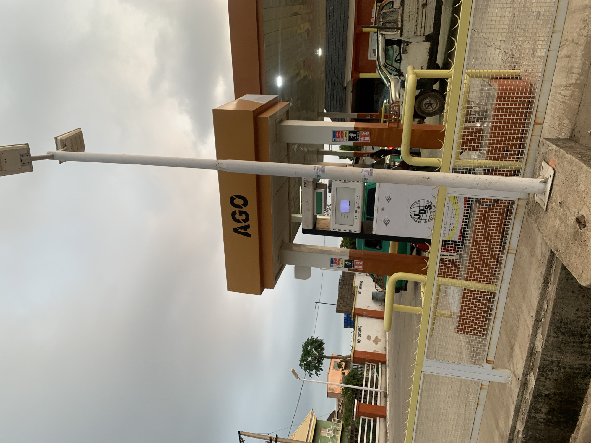 The image shows a gas station in Gura Top, Nigeria. The gas station has a brown and white awning with the word "AGO" written on it. There is a gas pump with a digital display and various warning signs attached to it. A white pickup truck is parked under the awning. A yellow and white metal fence surrounds the gas pump. A tall white light post stands to the side of the fence. The sky is overcast. Buildings, fences, and a small tree are visible in the background.