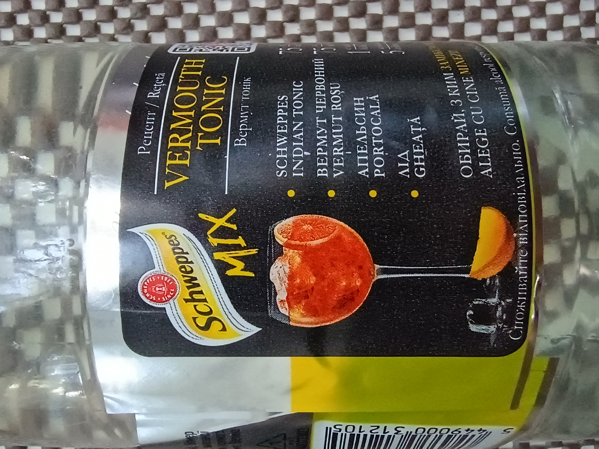 This is a close-up, eye-level, horizontal shot of a bottle of Schweppes Vermouth & Tonic. The bottle is the primary object, and its label is the main focus. The label is black with yellow and white text, and features a graphic of a cocktail glass with an orange garnish. The background is a textured brown and white checkered surface, likely a table or placemat.
The label displays a variety of text in different languages, including English, Romanian, and what appears to be Ukrainian. Prominent text includes "VERMOUTH TONIC," "SCHWEPPES," and "INDIAN TONIC." Other phrases like "Рецепт / Rețetă" (Recipe), "Вермут тонік" (Vermouth tonic), "ВЕРМУТ ЧЕРВОНИЙ" (Red Vermouth), "VERMUT ROŞU," "АПЕЛЬСИН" (Orange), "PORTOCALĂ," "ЛІД" (Ice), "GHEAŢĂ," and "ОБИРАЙ, З КИМ ЗАМІКА" (Choose who you mix with) are visible. There's also a barcode at the bottom with the number "5 449000 312105." The text "Споживайте відповідально. Consumă alcool re" is a responsible drinking message, translated into Ukrainian and Romanian.
The lighting appears to be ambient, with reflections on the glass bottle indicating it's indoors. There are no people visible in the shot. The context of Zaporizhzhia, Ukraine, suggests the location where the photograph was taken. The overall impression is of a product shot, highlighting the branding and ingredients of the beverage.