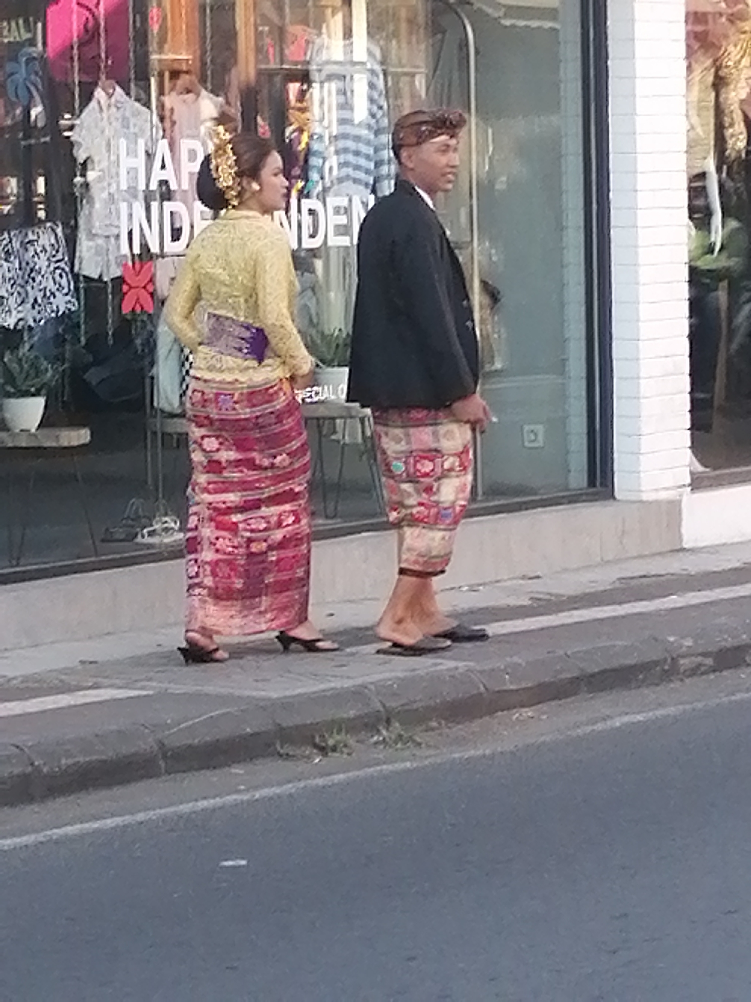 In front of a shop window displaying clothing and signage, two individuals are standing on a sidewalk. The individual on the left is a woman wearing a yellow blouse and a long, patterned red skirt, with her hair styled up with an ornate gold accessory. The individual on the right is a man wearing a black jacket, a patterned sarong, and a patterned head covering. He is holding a cigarette. Behind them, the shop window reflects the street and other people. The environment appears to be outdoors, with a paved street and sidewalk in the foreground. The shop appears to be located in a commercial area, possibly a tourist destination given the attire of the individuals. The signage visible in the shop window includes the words "HAPPY INDEPENDENCE". The overall scene suggests a daytime setting.
