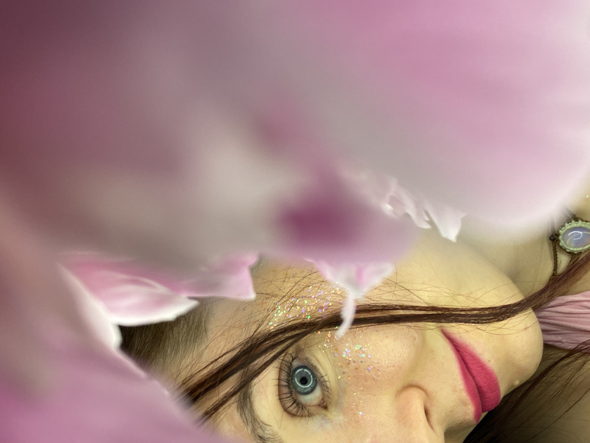 A close-up, artistic portrait features the partially obscured face of a person, likely female, framed by large, soft pink flower petals. The subject's head is tilted, with their face angled towards the viewer. Their striking blue-green eye, framed by dark eyelashes and eyebrows, looks directly at the camera. Glitter, predominantly in iridescent pinks and golds, is sprinkled on their forehead and cheekbone, adding a whimsical touch. Long, dark brown hair falls across their face and shoulders. Their lips are painted with vibrant, matte pink lipstick.
On the right side of the frame, near the subject's ear, a decorative earring or pendant with a pale, possibly opalescent, stone is visible, attached by a chain. Soft pink fabric, possibly part of clothing or a backdrop, is also visible. The foreground and background are largely blurred into soft gradients of pink, white, and purple, indicating a shallow depth of field and emphasizing the floral elements that envelop the subject. The overall mood is ethereal and dreamlike. There is no visible text in the image. No specific time of day or weather can be determined from the image, and there are no direct location cues referencing Chaikovsky, Russia.