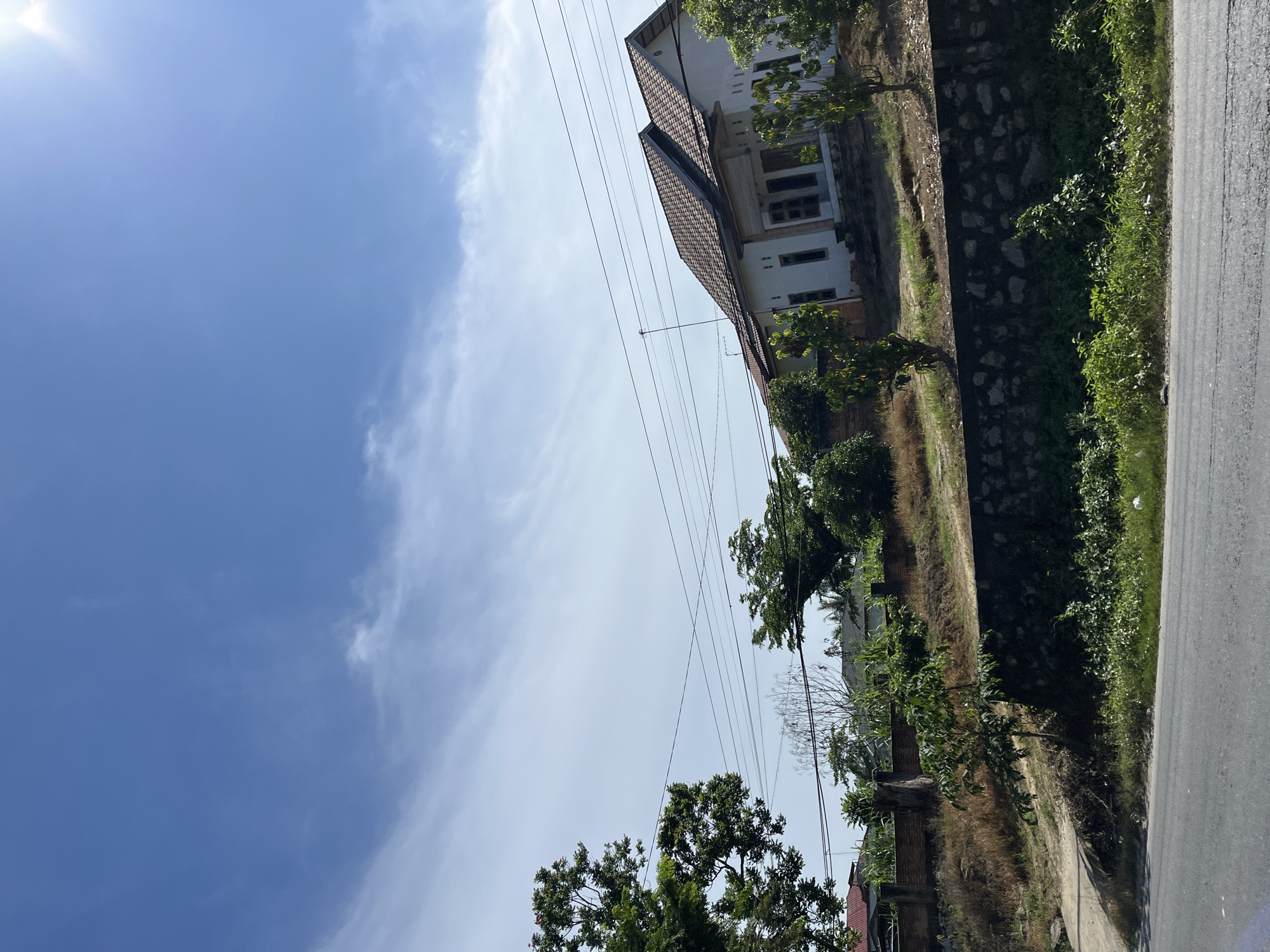 The media file shows a bright, sunny day in Bukitrata, Indonesia, with a clear blue sky and scattered white clouds. The image is oriented vertically, with the ground and buildings appearing on the right side and the sky on the left.
The main subjects are a building with a tiled roof and white walls, surrounded by greenery. There are several trees and bushes visible, suggesting a natural or semi-rural setting. A paved road runs along the right edge of the image, with a stone wall and a grassy embankment separating it from the property. Utility lines are strung across the scene, indicating the presence of infrastructure.
No people or specific activities are depicted. The overall impression is one of a peaceful day, likely during daylight hours given the bright sunlight and clear sky. The lush vegetation and the style of the building suggest a tropical or subtropical climate. There is no visible text in the image.