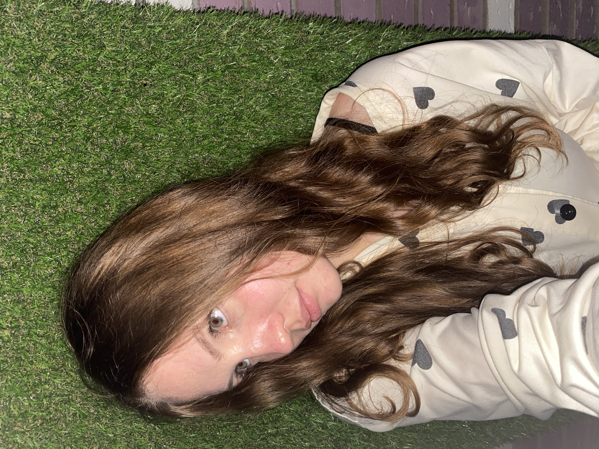 A woman lies on artificial turf. She has medium-length brown hair and is wearing a light-colored jacket with gray heart patterns. The scene includes the artificial turf, her hair, and her clothing. There is a brick wall in the background. There is no information to infer that this image was taken in the city of Izhevsk, Russia.