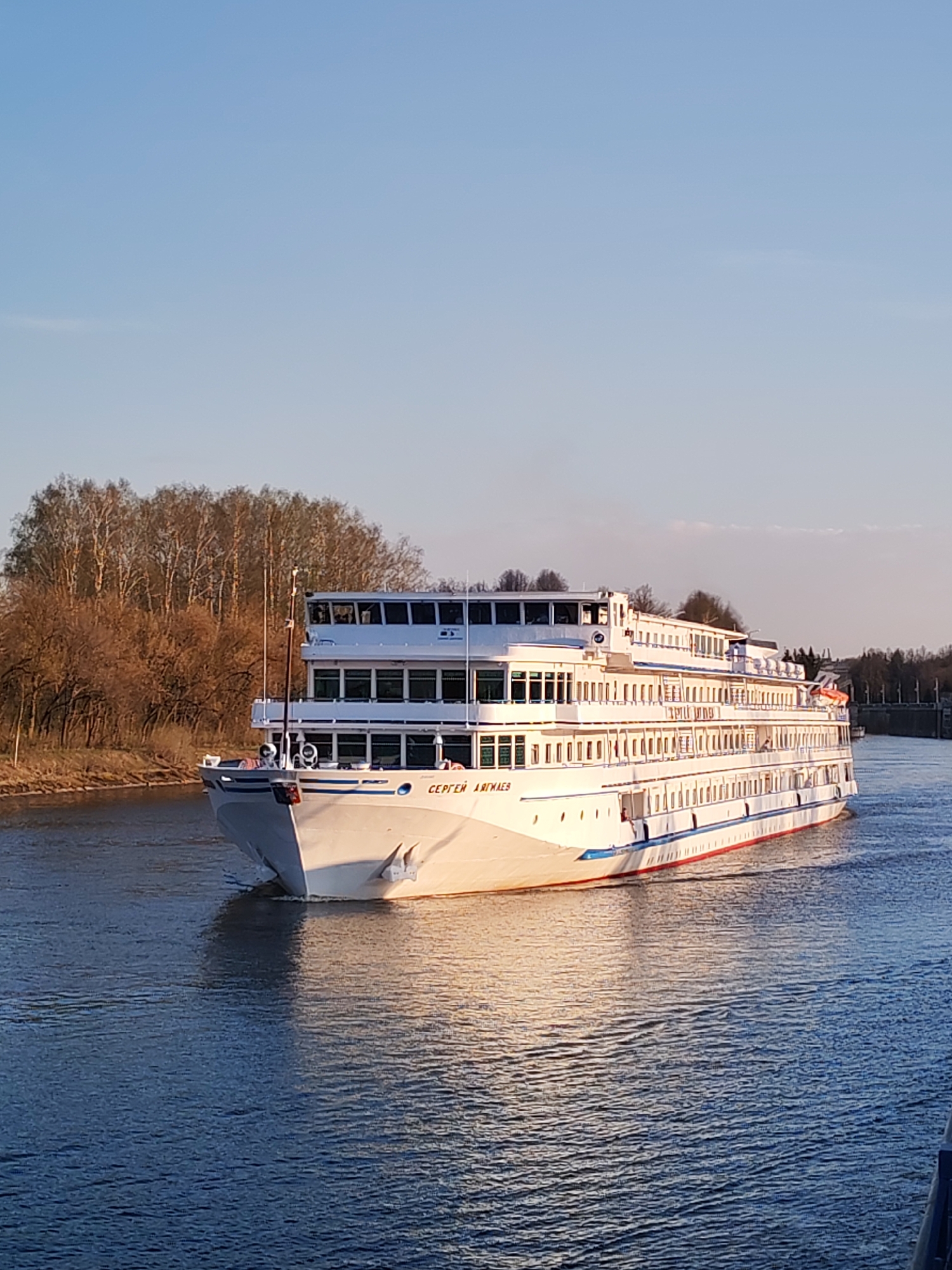 The image shows a river cruise ship, the "Sergey Dyagilev," sailing on a waterway in what appears to be Базарово, Russia. The ship is white with blue and red accents, and is several stories high. The surrounding environment consists of trees along the riverbank and a clear blue sky. The water is calm with reflections of the ship and sunlight.