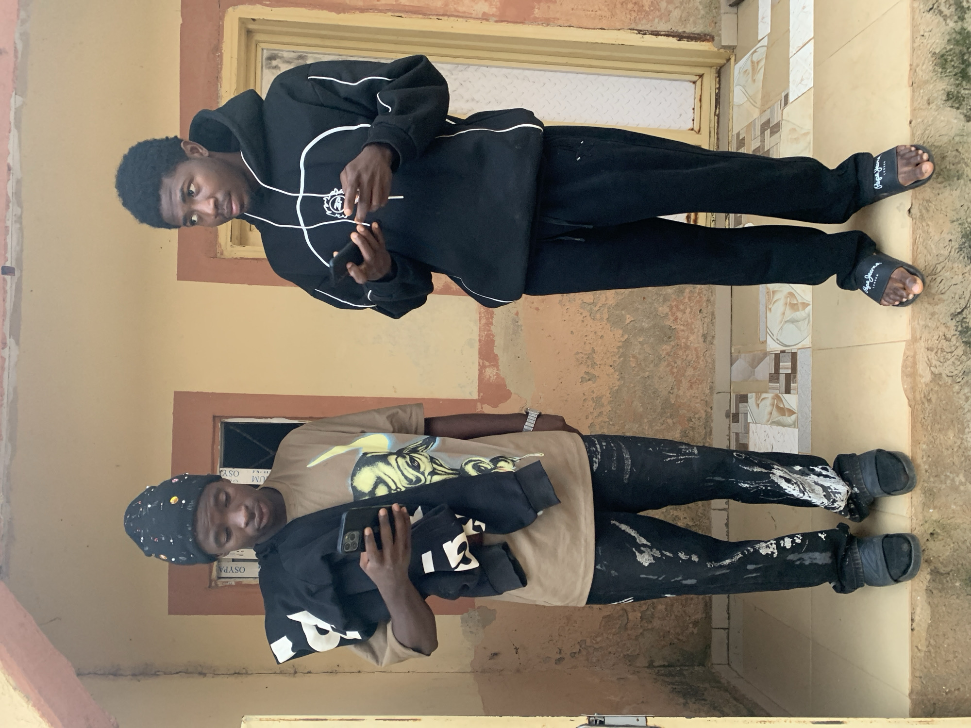Two young men stand in front of a building with a textured, yellow-ochre colored wall and a light brown door. The man on the left wears a black beanie adorned with colorful beads, a tan t-shirt with a graphic print, and dark pants splattered with white paint. He holds a dark-colored hoodie over his left arm and a smartphone in his right hand. He is wearing black slides. The man on the right wears a black hoodie with white piping and drawstrings, and black sweatpants. He holds a smartphone in his left hand and appears to be interacting with it. He is wearing black slides with white lettering. They are standing on a tiled porch. The environment appears to be outdoors, possibly in a residential area.