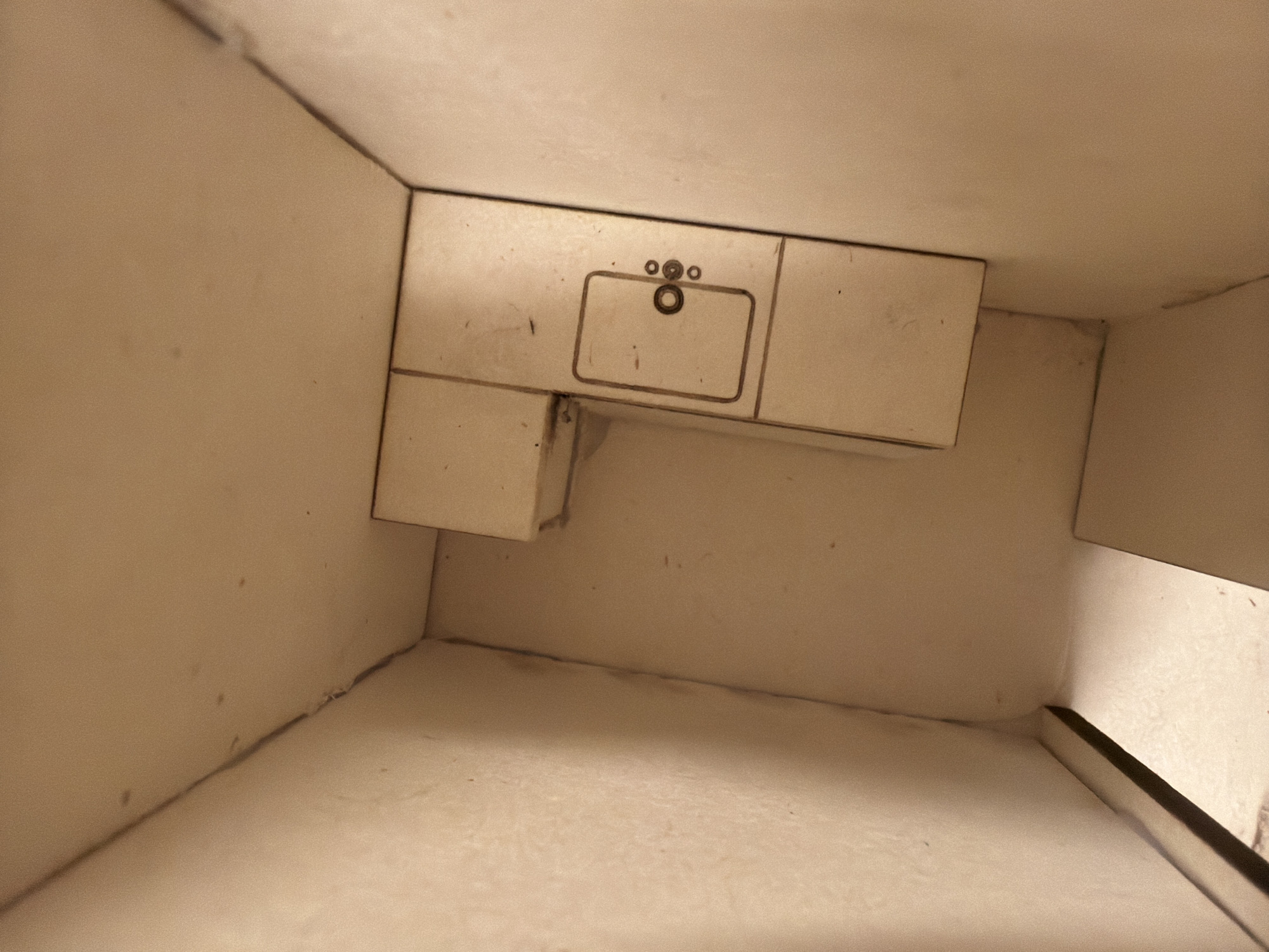 A top-down view of the interior of a miniature bathroom set constructed from pale, possibly unfinished wood. The set includes a sink and counter with three circular faucet fixtures, and a cabinet, all attached to the interior walls. The setting is likely an artistic rendering rather than a real bathroom. The location in the city of Milan, Italy is unknown.