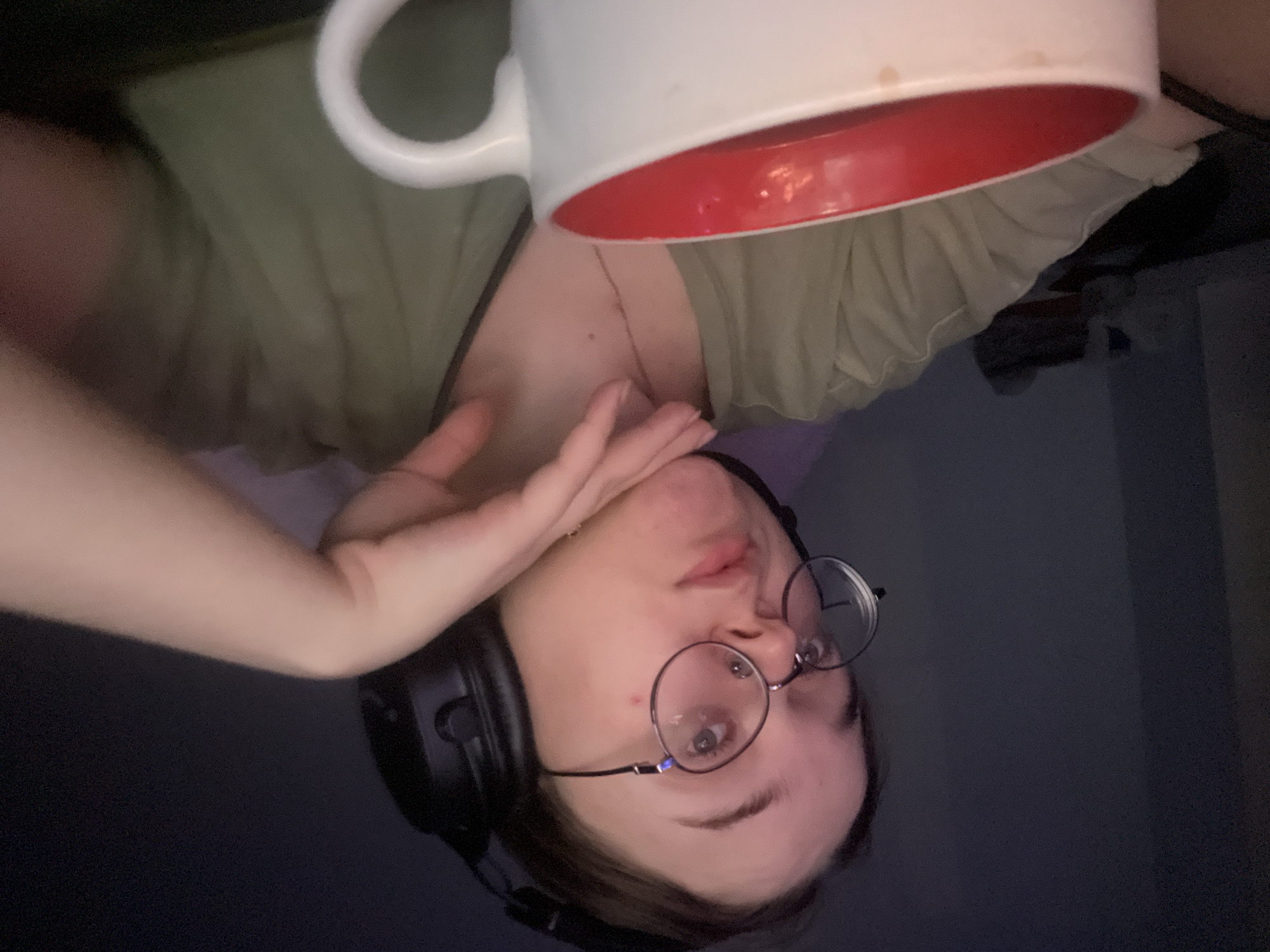 The image is an upside-down, close-up selfie of a person with fair skin, wearing round glasses, and black headphones. The person's face is in focus, showing one visible eye, eyebrow, and part of the mouth. The person's left hand is raised towards their face. They are wearing a light green shirt. Above the person is a large white mug with a red interior, which appears to contain liquid. The background is dark and indistinct. The location of the city of Pushchino, Russia, cannot be determined from the image.
