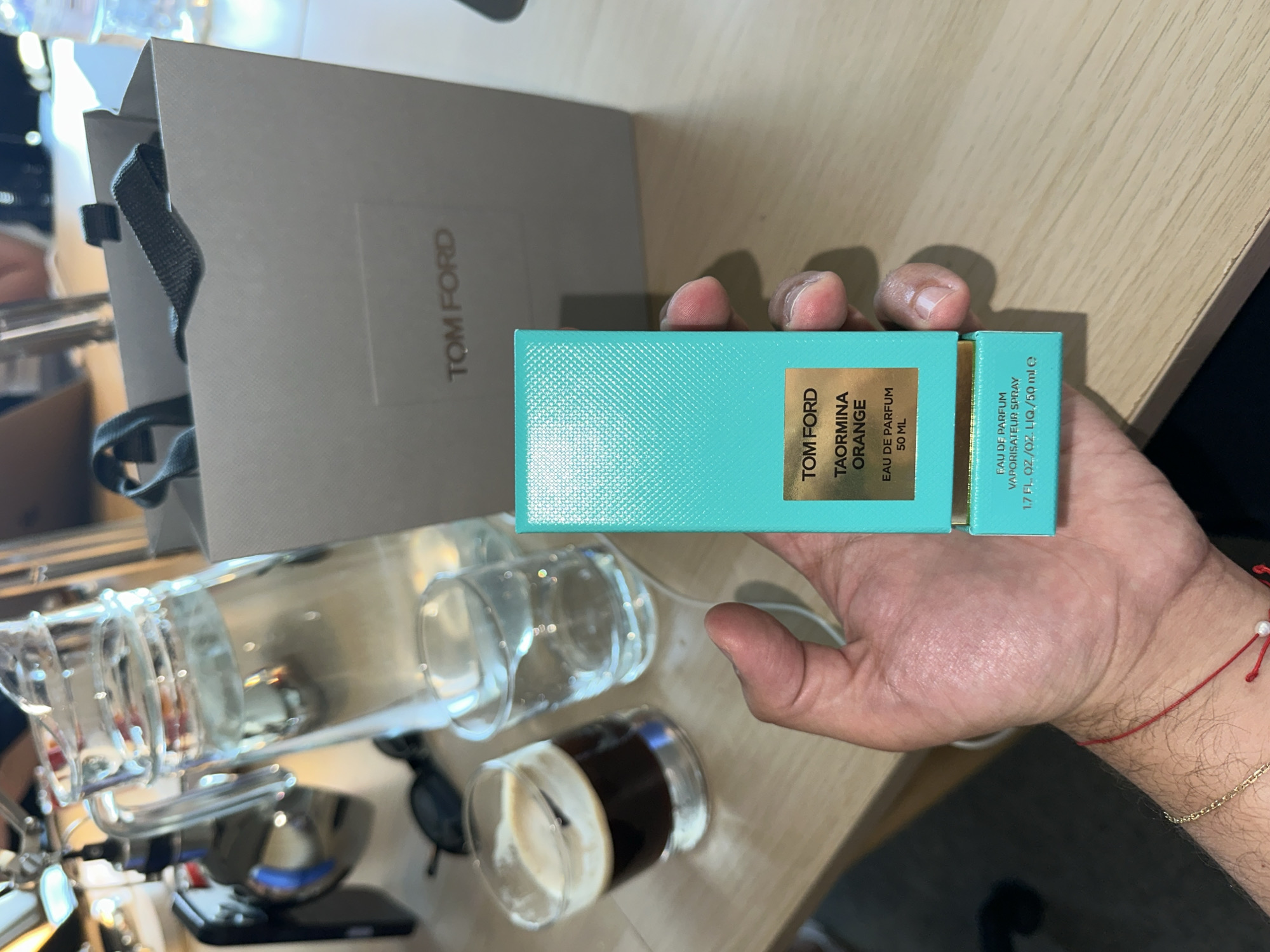 Here is a detailed description of the image:
A hand holds a turquoise Tom Ford Taormina Orange perfume box, next to a Tom Ford shopping bag. The box indicates "Eau de Parfum 50 ML." In the background, there is what appears to be a coffee cup, several glasses, sunglasses, and a smartphone. The setting seems to be a cafe or a similar indoor environment. The exact city location is unclear from the provided image.