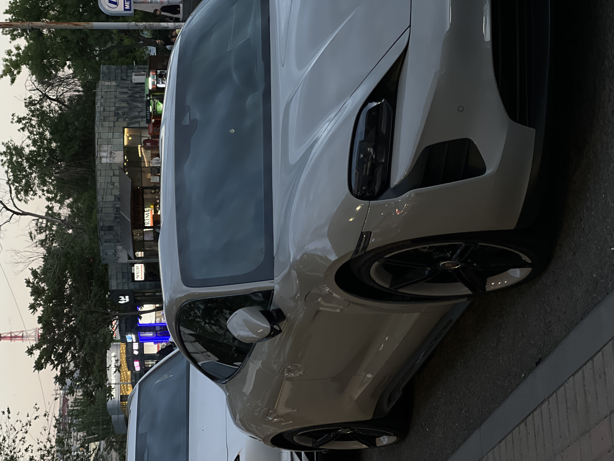 The image displays an urban street scene in Yerevan, Armenia, captured during what appears to be late afternoon or early evening, with soft, ambient light and some artificial illuminations. A modern, sleek, light-colored (likely white or very light gray) car, possibly a Porsche Taycan given its distinctive design, is the central focus, parked on the side of an asphalt road. Its front-right section is prominently featured, highlighting the unique LED headlights, sculpted fender, and a detailed front bumper with air intakes and a visible parking sensor. Black multi-spoke wheels are also visible.
Behind the main vehicle, another light-colored car is partially seen. The background features a row of commercial buildings, some with stone-block facades and various illuminated signs. Visible text includes "TELCELL" on a green sign and Armenian script, such as "ԻՄԻ" (IMI), on other storefronts. Lush green trees line the street, contributing to the urban landscape. In the upper right background, a "METRO" sign on a utility pole indicates a subway station nearby, with a few blurry figures of people discernible. The overall scene depicts a parked car in a city environment.