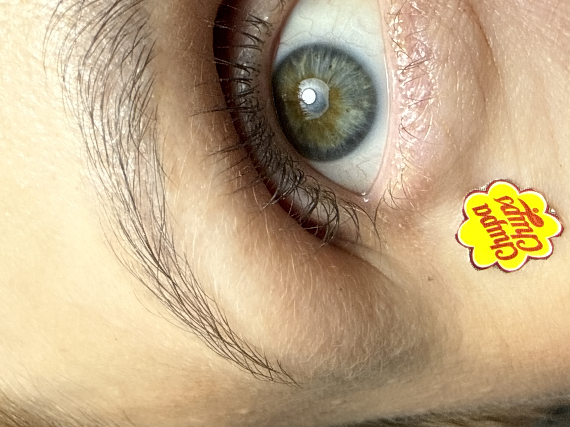 A close-up image focuses on a human eye with light green and yellow irises. The eye is surrounded by dark eyelashes and an eyebrow. Adhered to the skin below the eye is a yellow Chupa Chups brand sticker with red lettering. The skin appears smooth and fair-toned. No specific location in Chaikovsky, Russia, is discernible from this image.