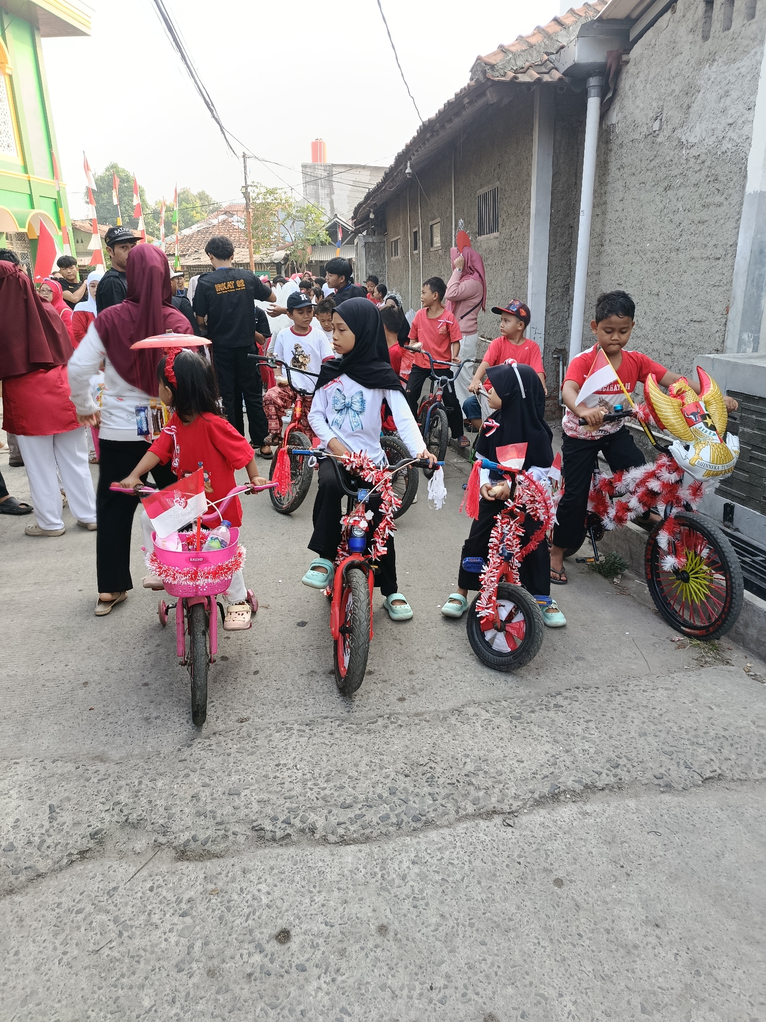 In Balaraja, Indonesia, a group of children are participating in a festive parade on bicycles decorated with red and white streamers, reflecting national colors. Several children are riding small bicycles adorned with Indonesian flags. One child is riding a bicycle with a large Garuda Pancasila emblem on the front. The parade is taking place on a paved street in a residential area, with houses visible in the background. The sky is overcast.