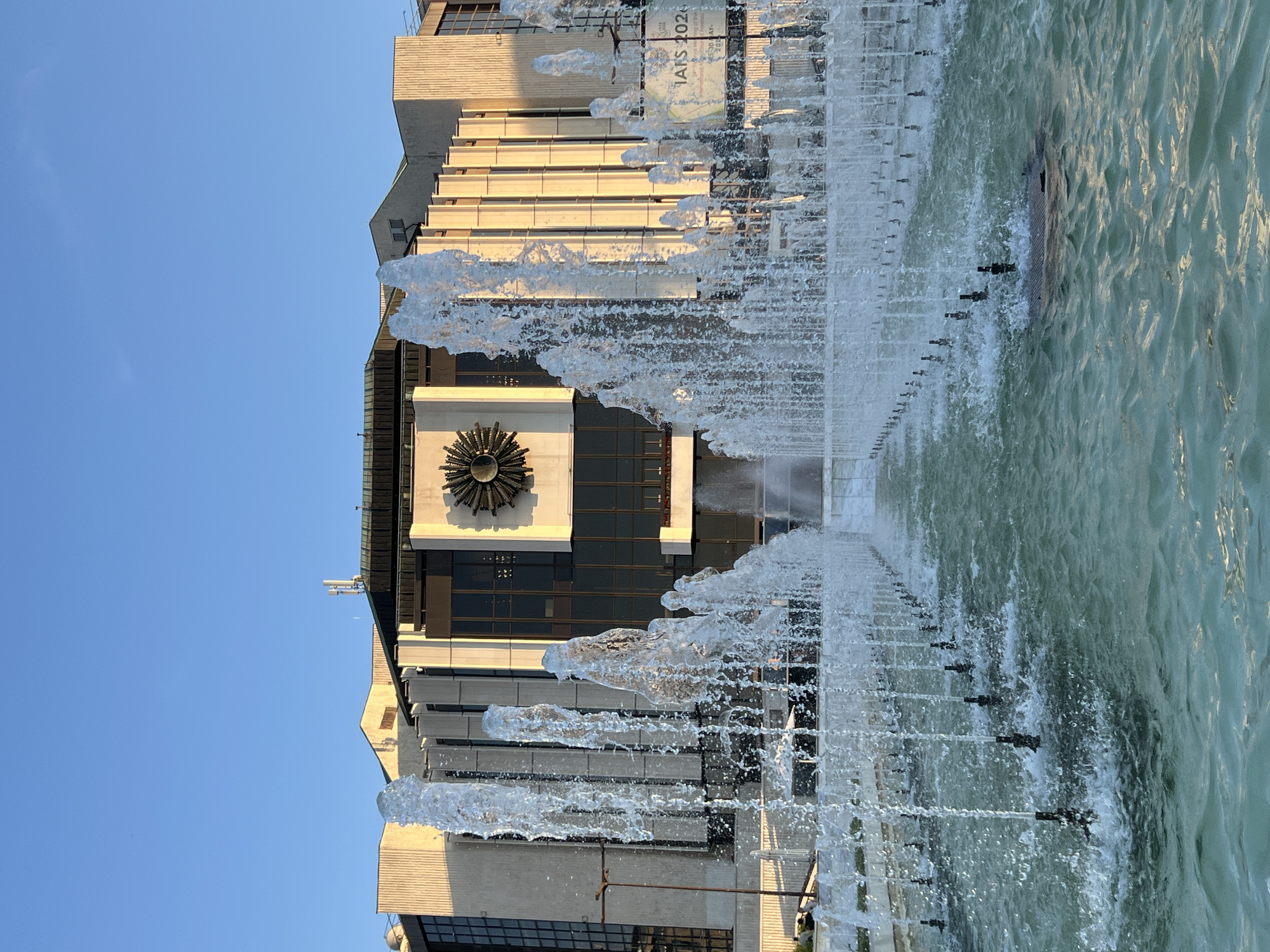 The image shows a large fountain in front of a building. The fountain has multiple jets of water spraying upwards, creating a misty effect. The building behind the fountain is modern in design, with a concrete facade and large glass windows. There is a circular sun-like emblem mounted on the building. A banner is visible on the right side of the building with the text "IANS 2020". The sky is clear and blue. The lighting suggests it is either early morning or late afternoon.