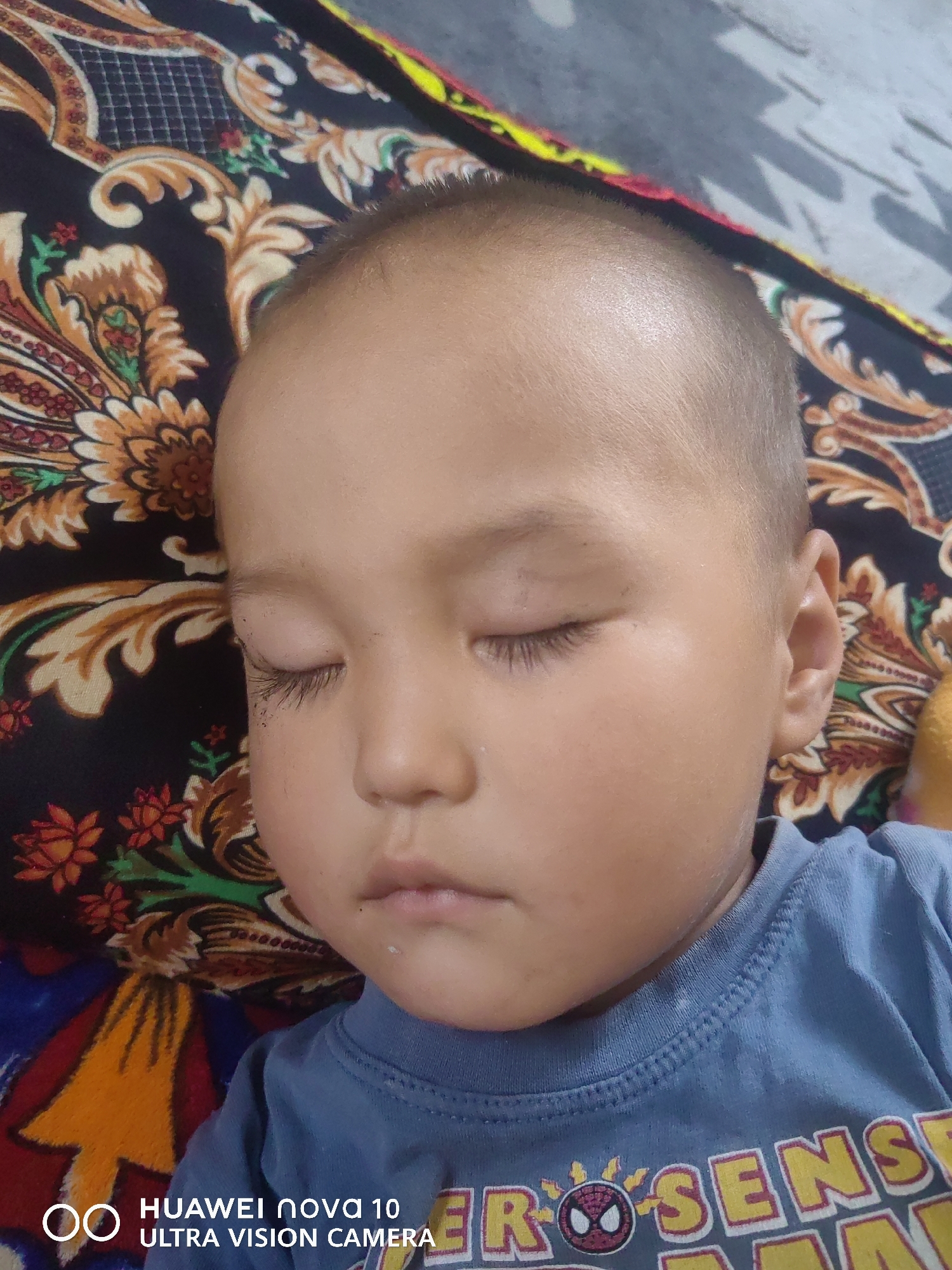 A close-up, eye-level shot shows a young child sleeping on a patterned blanket. The child, with fair skin and dark hair, is positioned on their back with their eyes closed. They are wearing a blue t-shirt with a yellow graphic that appears to be a cartoon character. The background is a dark, richly patterned fabric with floral motifs in orange, yellow, and red. The image also includes a watermark in the lower left corner that reads "HUAWEI nova 10 ULTRA VISION CAMERA". This image was taken in Jetim-Dobo, Kyrgyzstan.