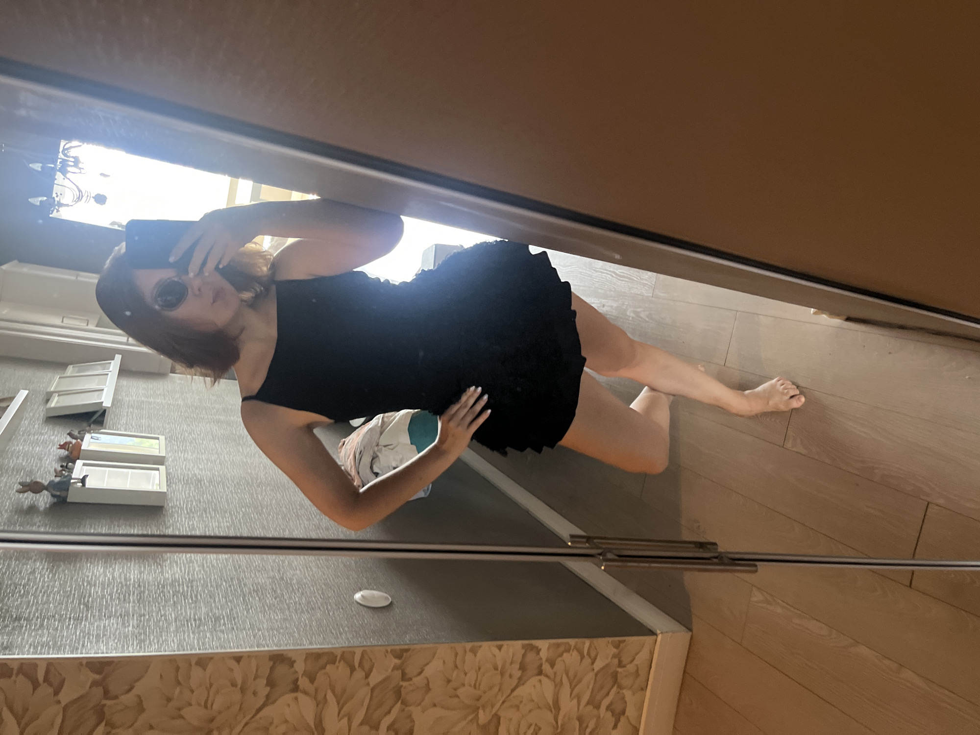 A young woman is standing in a room, taking a mirror selfie. She is wearing a short black dress and sunglasses, and is holding a smartphone in her right hand. Her left hand is on her hip. The room appears to be a bedroom or dressing room, with a large wardrobe with mirrored doors on the left side of the frame. The walls are decorated with a floral wallpaper on the left and a textured gray material on the right. Shelves with decorative items are visible on the right wall. The floor is made of light-colored wooden planks. The lighting suggests it is daytime, with light coming from a window or lamp reflected in the mirror. The scene is indoors, likely in an apartment. There is no visible indication of the specific location in Krasnoyarsk, Russia.
