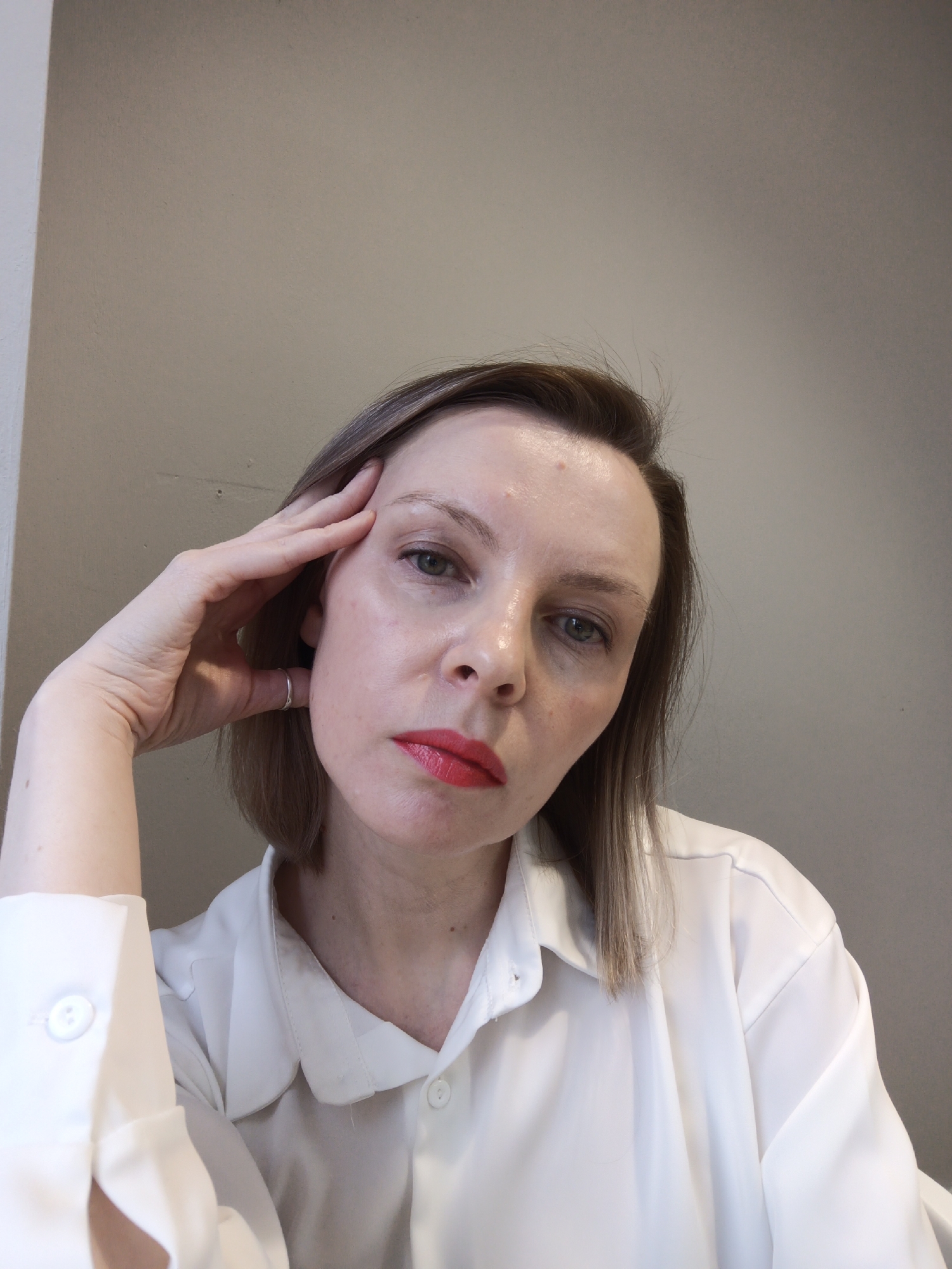 A Caucasian woman with short, light brown hair is seen from a low-angle perspective. Her face is tilted slightly downwards, and she is resting her right hand against her forehead, with a simple silver ring visible on her ring finger. She has fair skin, light eyes, and is wearing red lipstick. The woman is wearing a white, collared button-down shirt. The background is a plain beige wall. There is no indication that the image was taken in Chaikovsky, Russia.