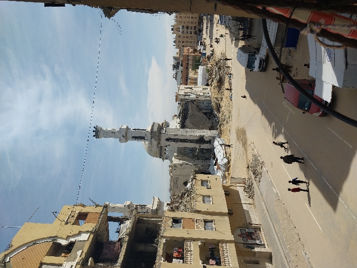 The image depicts a devastated urban street scene in Khan Yunis, Palestinian Territory, under a partly cloudy blue sky. In the center, a white mosque, identified as Al Farouk Mosque, stands with its tall minaret and dome largely intact, though surrounding structures are reduced to rubble. To the left, a multi-story, light brown building exhibits severe damage, with large sections of walls missing, exposing interiors and balconies where laundry hangs. Extensive piles of concrete and building debris litter the ground, particularly around the mosque and along the street edges.
A paved road with faded white markings runs through the foreground and midground. Several individuals are present: three children walk together on the left side of the street, followed by an adult male. Another child walks further ahead. In the background, near the mosque and rubble, multiple small figures are visible, some appearing to be children, moving through the debris-strewn area. Vehicles include a red sedan parked on the right in the foreground and a white panel van parked further down the street on the right. Electrical or communication wires are stretched horizontally across the upper mid-ground. The foreground right includes a partial view of a dark coiled object and a white tarp structure, indicating the vantage point is from an elevated position. The scene conveys widespread destruction with elements of ongoing human activity amidst the ruins.