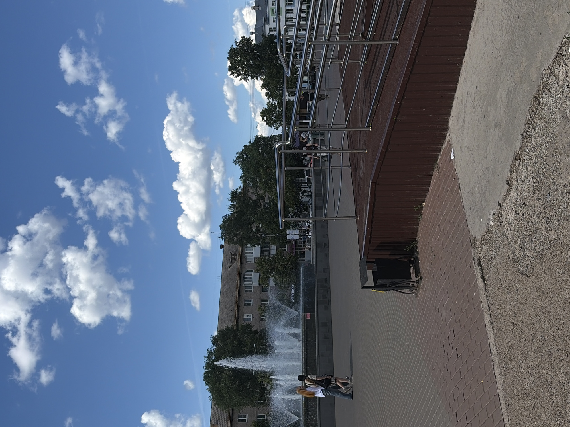 The image shows a sunny day with a bright blue sky and scattered white clouds. In the foreground, a paved area made of brick-like stones leads to a ramp with chrome railings. A dark brown wooden fence runs along the base of the ramp. To the left, a black trash can with ornate legs stands on the pavement.
In the middle ground, a large fountain with several jets of water is in operation. Behind the fountain, a multi-story building with a grey facade and numerous windows is visible, flanked by mature green trees. Two young women are walking side-by-side on the paved area in front of the fountain, one wearing a white shirt and jeans, the other a black top and shorts.
Further in the background, other buildings and more trees can be seen. Some people are visible walking in the distance near the buildings. The overall impression is of an outdoor public space in an urban environment.