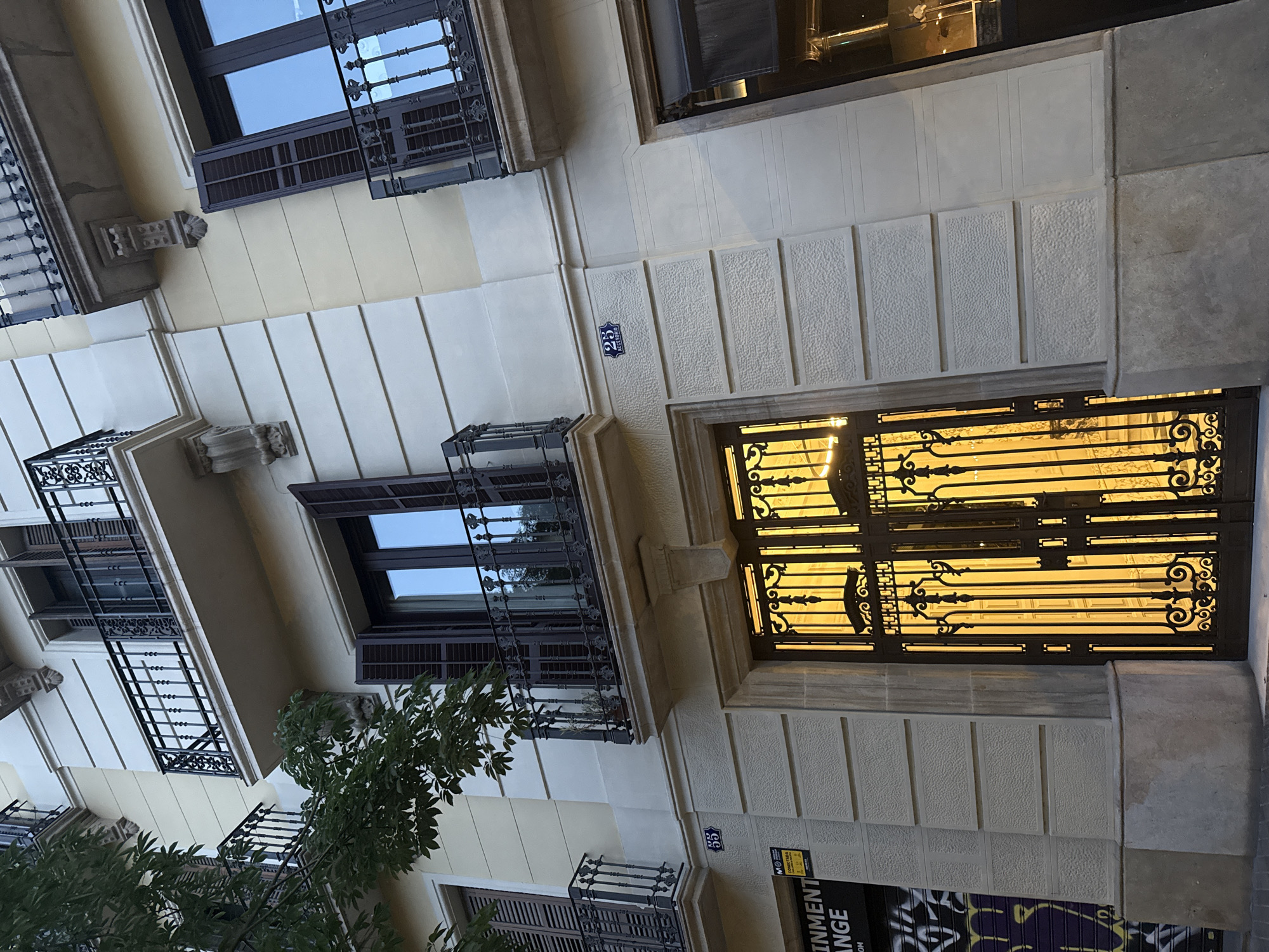 This is a medium-shot, eye-level, vertical photograph of the facade of a building in Barcelona, Spain, with a tree partially obscuring the left side. The building has light yellow and cream-colored walls with ornate black wrought-iron balconies on multiple floors. The balconies have decorative scrollwork and are supported by stone corbels. Two windows with dark brown shutters are visible on the upper floors, one of which is open. Below the balconies, the facade is decorated with horizontal lines and textured panels.
At street level, a large, ornate black wrought-iron gate covers an entryway. The gate is intricately designed with floral motifs and vertical bars, and a warm golden light emanates from behind it, suggesting interior lighting. To the left of the gate, a section of the wall displays a yellow sign with black text that reads "MONEY EXCHANGE". Below this sign, graffiti in purple and black spray paint is visible on a rolled-down metal shutter.
A small blue and white street sign with the number "25" and the word "ACCESORIO" is affixed to the wall above the gate. The building's base is a textured stone foundation. The overall scene depicts a well-maintained urban building with elements of classic European architecture.