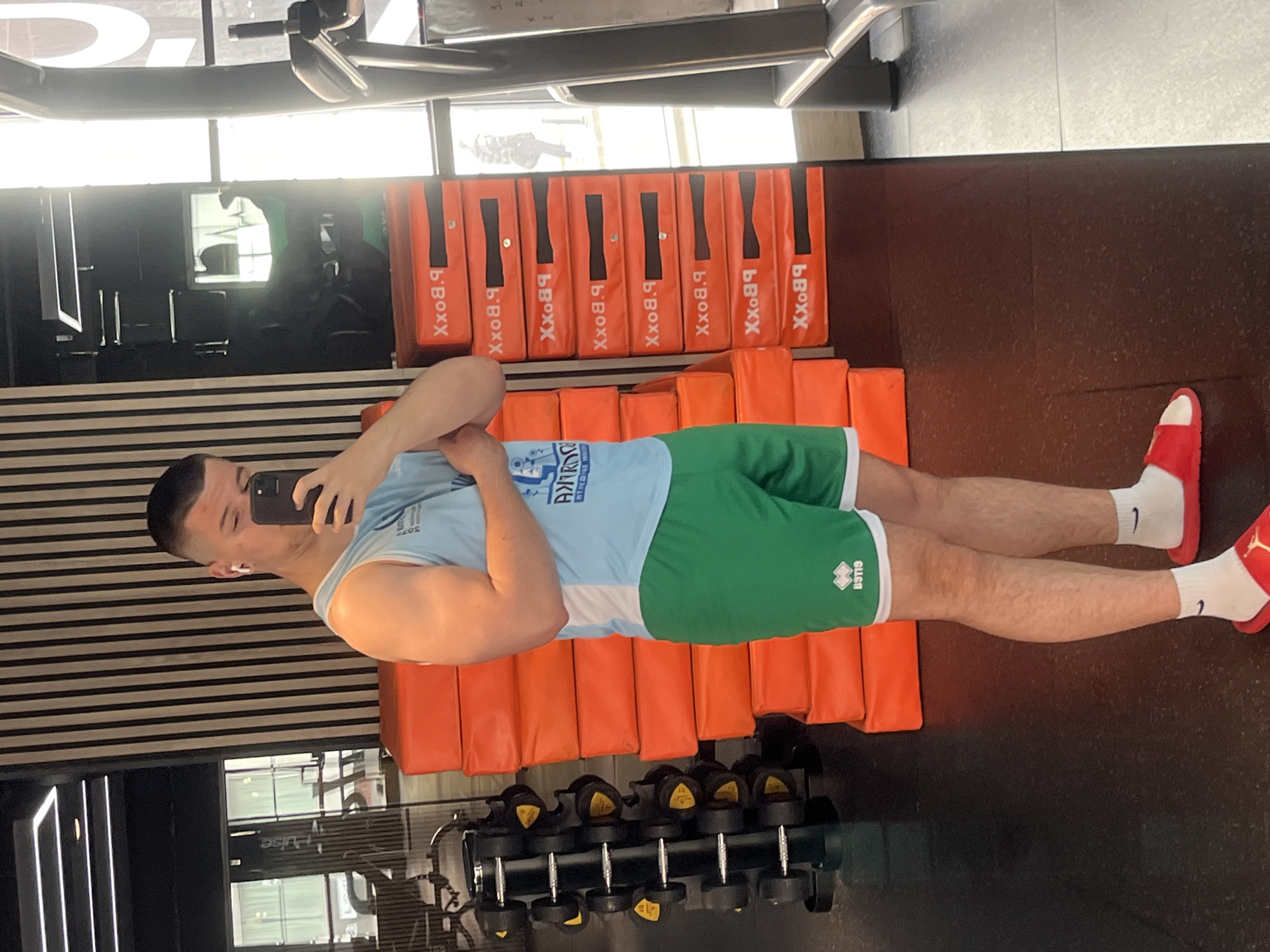 A man is in a gym. He is wearing a light blue and white tank top, green shorts, white socks, and red sandals. He is holding a black smartphone up to his face as if taking a selfie. Behind him is a row of orange pads with "B.Boxx" printed on them. Behind the pads is a black and white striped wall. In the foreground, there is a rack of dumbbells. The gym appears to be located in Sofia, Bulgaria, based on the ANTIPOV/RVD SOFIA logo on the man's tank top.