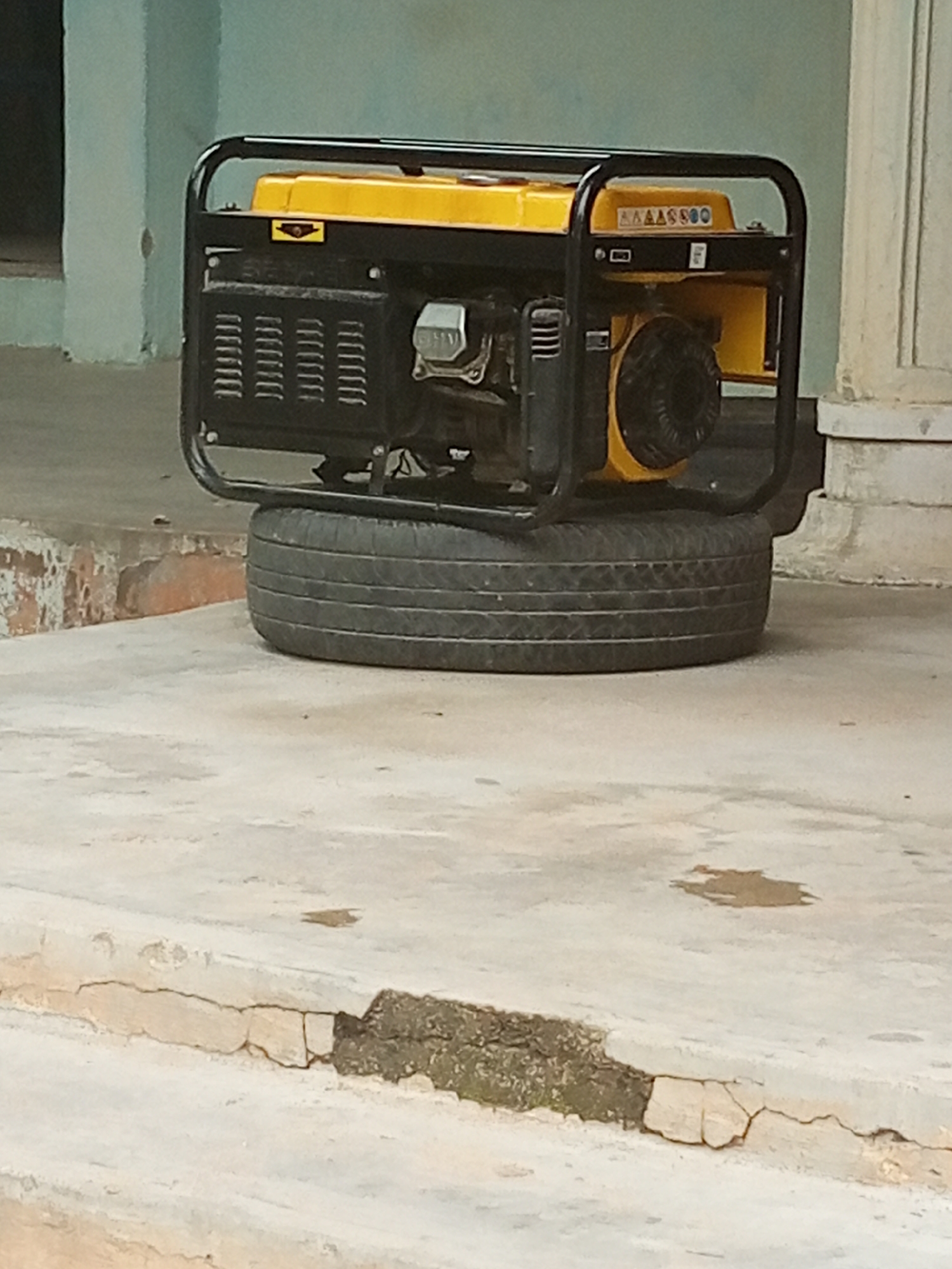 A yellow and black portable generator is positioned on top of a tire, which itself rests on a concrete surface. The generator is supported by a black metal frame. Visible on the generator is a yellow panel with some black markings. The background shows a light blue wall and a portion of a white pillar. The scene appears to be outdoors, possibly in a yard or on a patio, given the concrete ground and the outdoor setting implied by the lighting. There is no activity taking place; the generator is stationary. The lighting suggests it is daytime, though the exact time is not discernible. There are no people visible in the image. The concrete shows some wear and tear, with cracks and discoloration, suggesting an outdoor environment exposed to the elements. The overall impression is of a functional object placed in a somewhat utilitarian setting. There is no discernible text that can be read clearly.