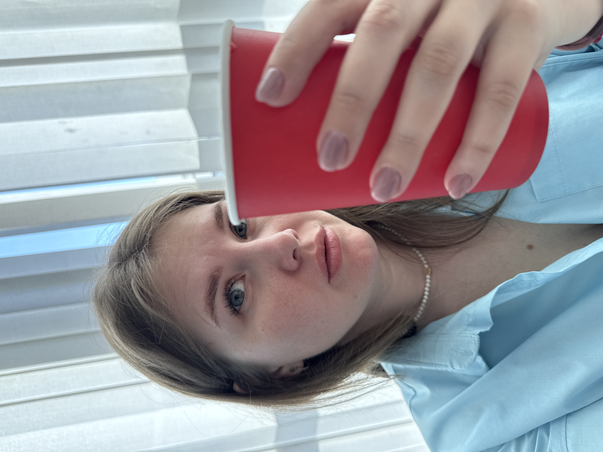 A low-angle, portrait-style photograph captures a young woman looking directly at the viewer. Her light brown hair falls around her shoulders, and her face, with light skin and blue-grey eyes, holds a neutral to slightly serious expression. She wears a light blue collared shirt and a delicate pearl necklace. Her right hand, featuring light pink nail polish, is prominently positioned in the upper right foreground, holding a red disposable cup. The background is dominated by white venetian blinds, indicating an indoor setting with bright, natural light filtering through, suggesting it is daytime. There is no visible text in the image.