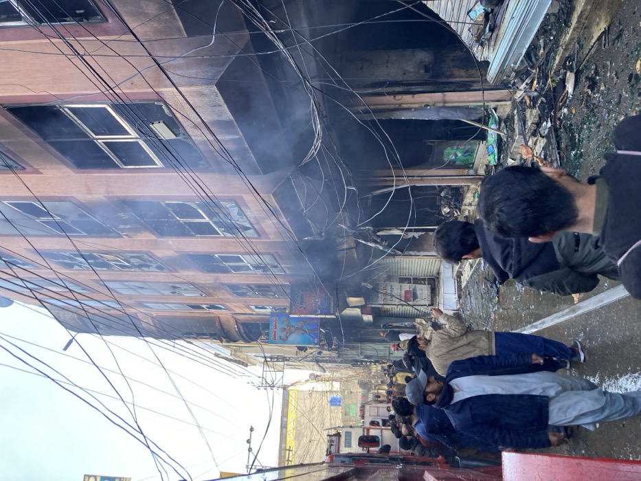 The image depicts the aftermath of a fire on a narrow urban street in Quetta City Tehsil, Pakistan. A multi-story building, at least four floors tall, occupies the right side of the frame, exhibiting extensive fire damage. Its facade, originally a light brown, is heavily blackened by soot and charring, particularly on the lower levels and around window frames. Numerous windows on all visible floors are broken, shattered, or missing glass panes. Smoke is actively emanating from the lower floors and ground level.
The street in front of the damaged building is wet and covered with debris, including shattered glass, charred materials, and twisted metal. An intricate network of overhead electrical and utility wires crisscrosses the scene, with many wires appearing damaged, melted, or dislodged due to the fire. Shop fronts on the ground floor are severely burned and gutted, with one visible sign for "S.R.K Laptops" on a partially closed metal shutter. Another sign higher up features a muscular figure and Urdu text.
A crowd of approximately 30-40 adult males is gathered on the street, observing the scene. Most are dressed in casual attire, including jackets and traditional Pakistani clothing. Some individuals are walking, while others stand still. One man in the midground holds a long pole or rod. In the background on the far left, two fire trucks, one red and one white, are partially visible, indicating an emergency response. The sky above is overcast or hazy white.