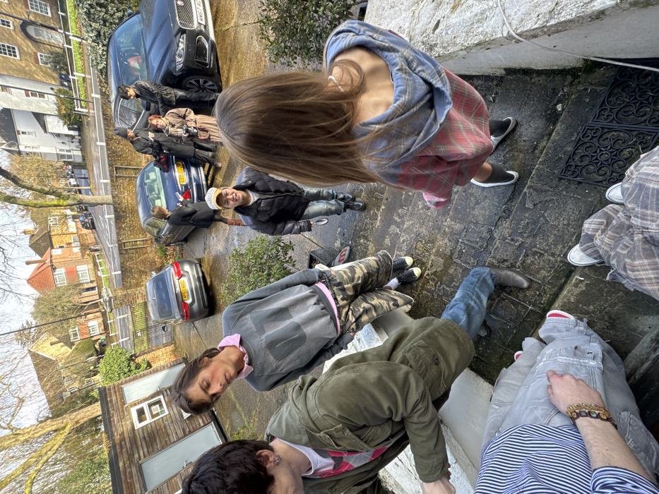 The image depicts a daytime scene in a residential area of London, United Kingdom, characterized by multi-story brick and stucco houses and deciduous trees without foliage, indicating a late autumn or winter setting. A paved driveway or street hosts three parked vehicles: a grey sedan with UK license plate WPE 6 XAO, a blue sedan with partially visible UK plate GC03 DPY, and a black Jaguar SUV displaying UK license plate WM67 ADJ.
At least ten individuals are present on a concrete path and stone steps leading down from an elevated position. In the lower foreground, a person is partially visible from the waist down, wearing grey pants, a striped shirt, a beaded bracelet on the left wrist, and red sneakers. Adjacent, another person's lower legs are visible, clad in plaid pants and white sneakers.
Mid-ground, on the steps, a male with dark hair, wearing a dark green jacket, blue jeans, and brown boots, appears to be descending. Standing on the paved area, a thin male with shoulder-length brown hair wears a pink collared shirt under a distressed grey sweatshirt, camouflage shorts, and black sneakers. Opposite, a female with long brown hair, viewed from behind, wears a red and blue plaid hoodie and black and white footwear. Near her, a male in a dark puffer jacket, light jeans, black boots, and a light-colored baseball cap smiles while holding a small object, looking towards the female in the plaid hoodie.
In the background, a group of four individuals stands near the parked cars. This group includes a female in a dark coat holding a phone, a female in a long brown coat holding a camera, a male in a dark jacket and beanie holding a phone, and another male in a dark jacket. A wooden shed with a window and door is situated on the left side of the frame. The ground exhibits patches of dampness, and a black decorative doormat rests on the steps.
