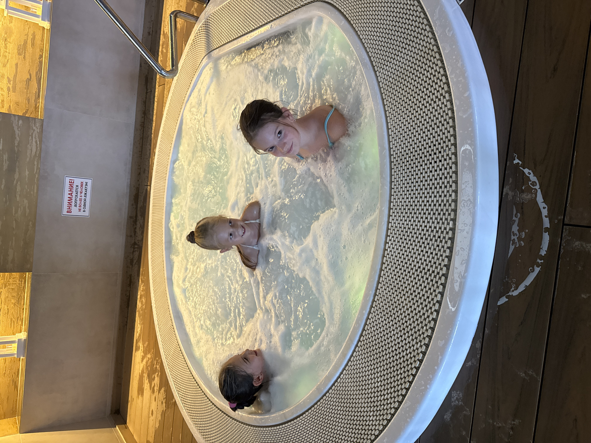 Three young girls are in a hot tub filled with bubbly water. The girl on the left, with dark hair pulled back in a bun, smiles at the camera. The girl in the middle, with blonde hair in a ponytail, also looks towards the camera. The girl on the right, with dark hair styled in a bun, smiles and looks at the camera. They are wearing swimsuits. The hot tub is round with a textured, light-colored rim and is set on a dark wooden floor that appears wet. In the background, a sign with Russian text is visible on a light-colored wall. The overall environment suggests an indoor pool or spa facility.