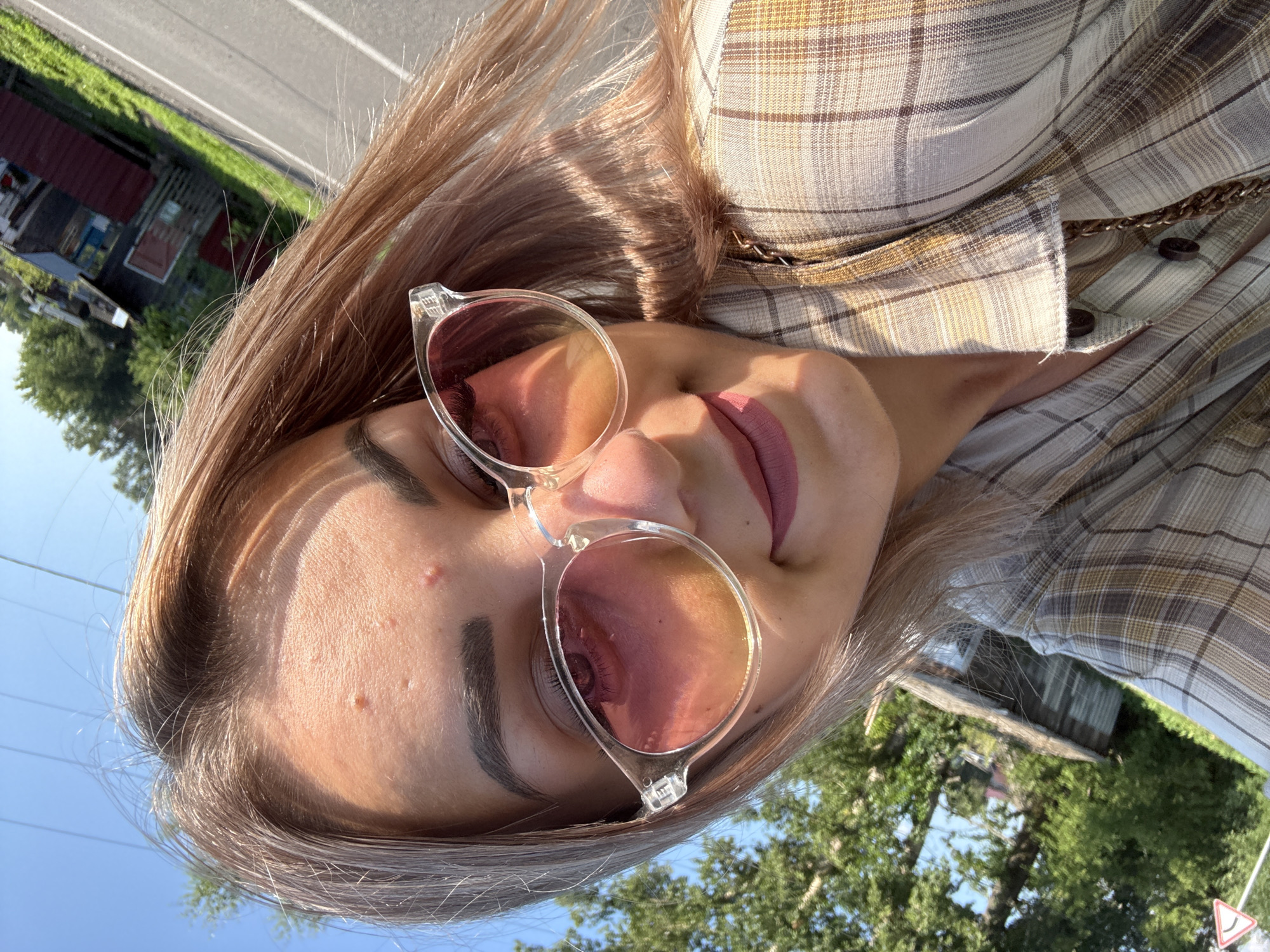 A young woman with blonde hair, wearing pink lipstick and clear-rimmed sunglasses with pink lenses, takes a selfie outdoors. She is wearing a plaid shirt and has a chain strap visible over her shoulder. Her head is tilted, and she is looking down. The background shows greenery, trees, a dirt road, a small building, and a clear blue sky. The lighting suggests it is daytime, possibly late afternoon. The location is the city of Krasnogorskoye, Russia.