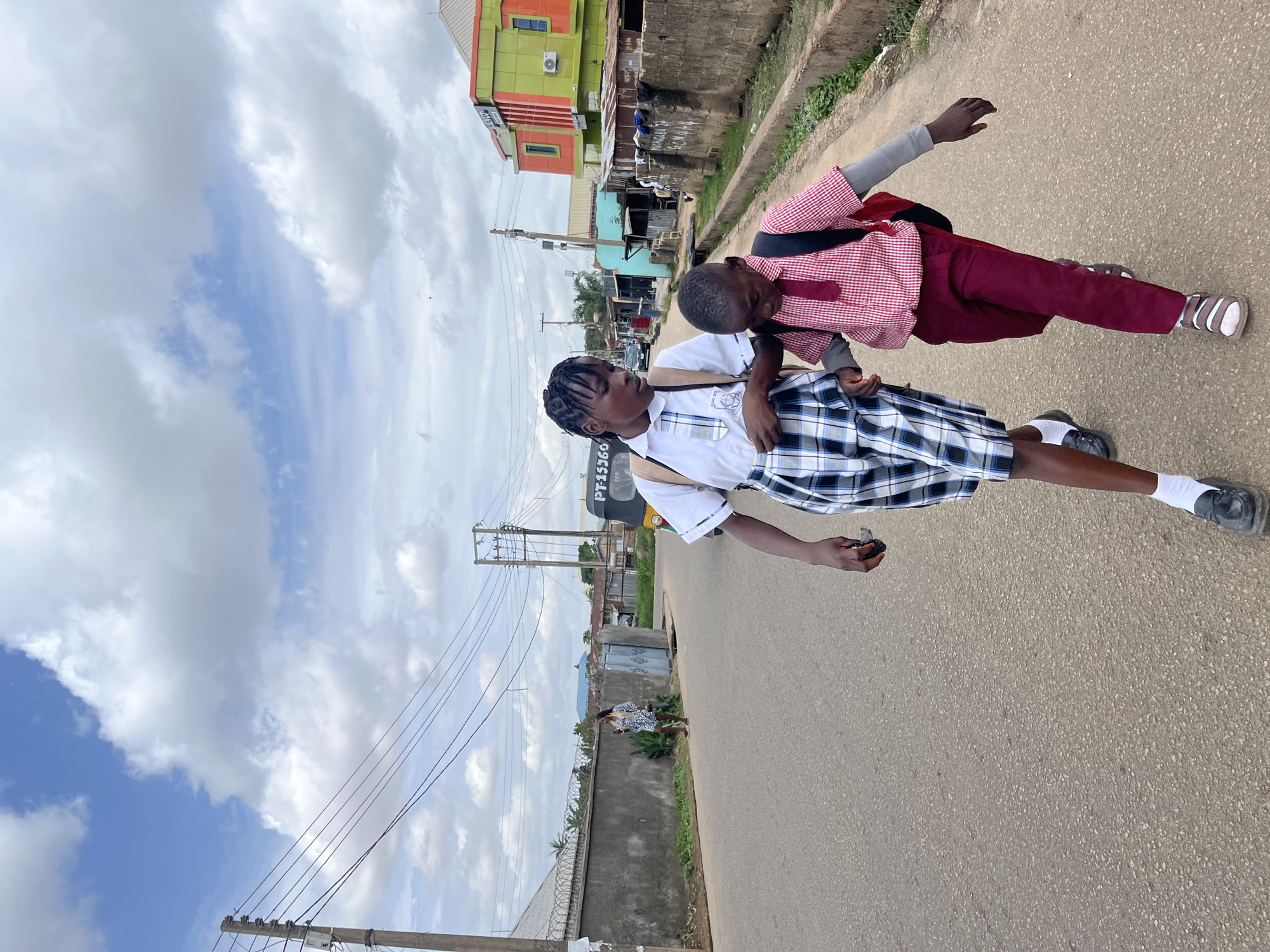Two young people, likely students based on their uniforms, are walking on a paved road in Jos, Nigeria. The person in the foreground wears a white short-sleeved shirt with a plaid tie and a plaid skirt. They have dark hair braided and are looking to the left. The second person, behind and slightly to the right, wears a red and white checkered shirt with a dark tie and maroon trousers. This person is looking down and to the left. Both individuals have backpacks. The scene is an outdoor setting with a cloudy sky. Buildings, some brightly colored, and utility poles line the side of the road. A partial view of a tuk-tuk is visible in the background. The time of day appears to be daytime, given the lighting. The overall mood seems casual, perhaps on the way to or from school. Visible text includes "PT-15360" on the side of the tuk-tuk.