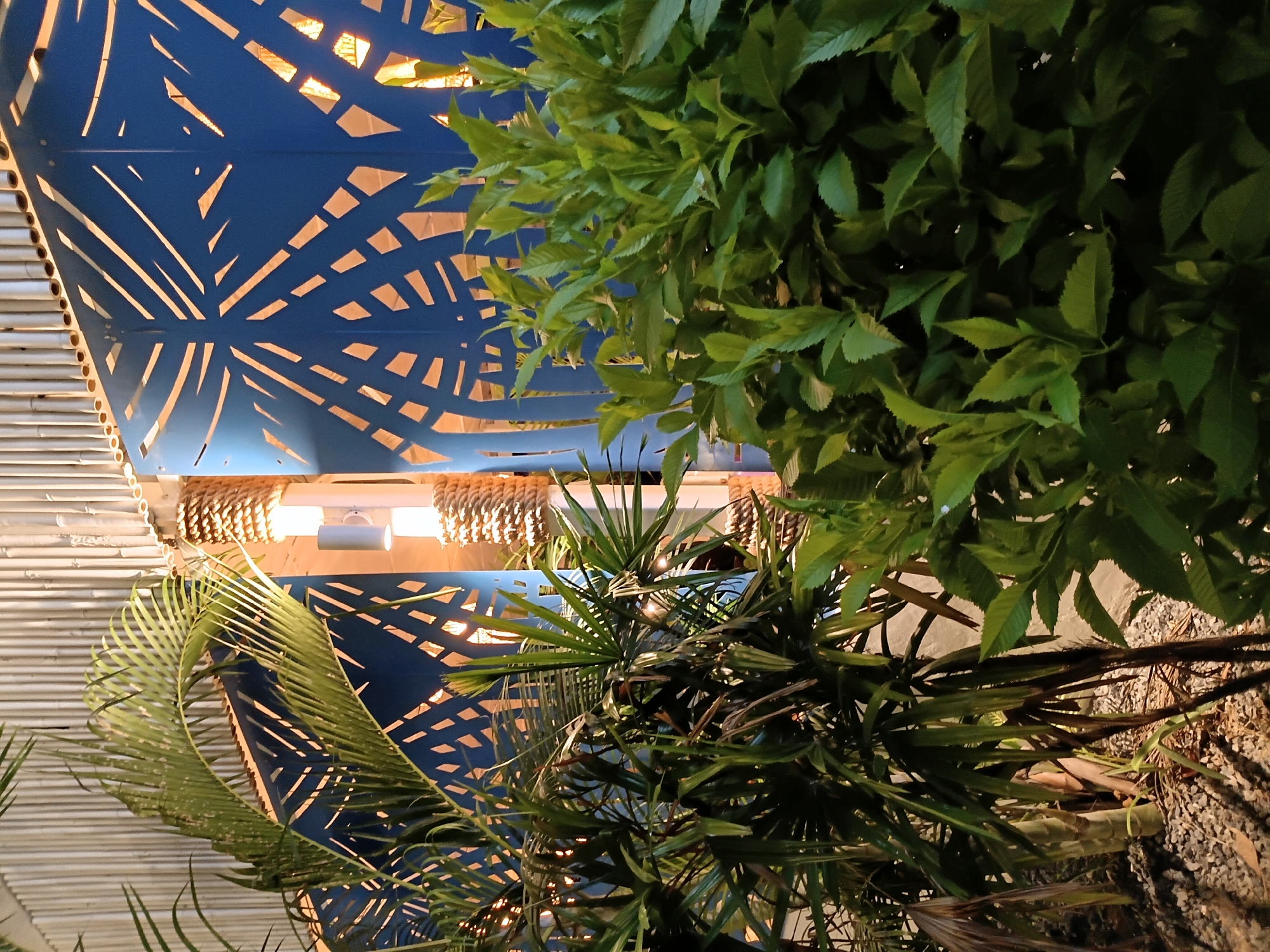 The image, likely taken at night, shows an outdoor space in Baan Sri Thanu, Thailand. The perspective is looking upwards towards a partially obscured ceiling structure. The ceiling consists of a blue, patterned metal panel with geometric cutouts allowing light to pass through. This panel is supported by a lighter-colored, possibly bamboo or reed, frame with hanging rope decorations. Lush green foliage, consisting of trees or bushes with various leaf types, dominates the right and lower portion of the frame, partially obscuring the structural details. The ground is visible in a small area at the bottom and appears to be covered with gravel or small stones. The illumination is artificial and creates a warm glow through the patterned ceiling. There are no people visible in the scene.