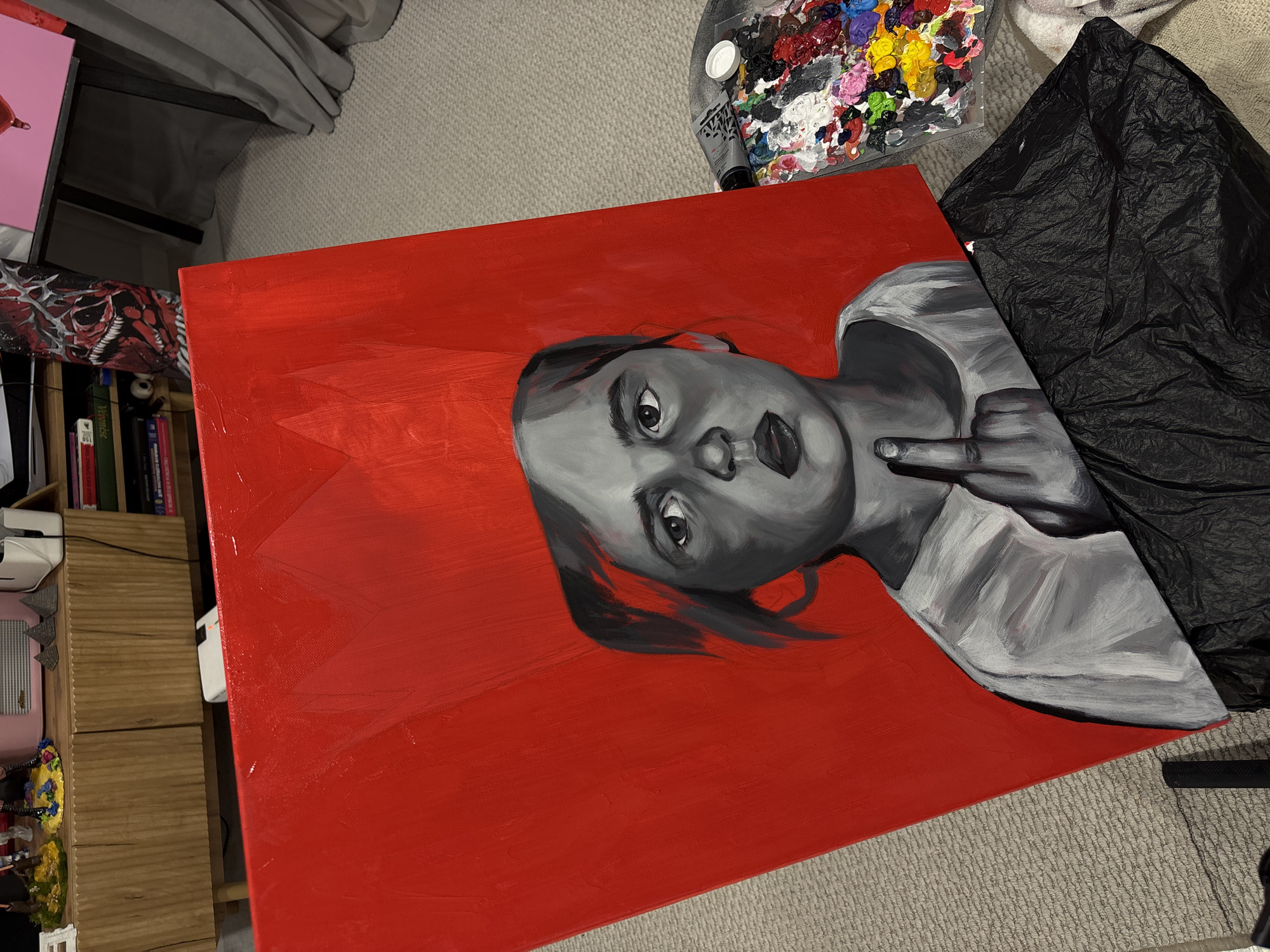 A rectangular canvas painting, measuring approximately 1.5 times wider than it is tall, is laid horizontally on a light grey carpeted floor in an indoor setting. The painting features a bold, graphic portrait of a young person, likely female, rendered in grayscale (black, white, and shades of grey) against a vibrant, solid red background. The figure looks directly at the viewer with wide, intense eyes and slightly parted lips. Her dark hair is styled, and she wears a light-colored top. Her right hand is raised, prominently displaying an extended middle finger.
Surrounding the painting, various objects suggest an artistic workspace. To the upper right, a plastic paint palette is visible, covered with numerous splotches of colorful paints, indicating recent use. Next to the palette, a tube of grey paint is present. A crumpled piece of black fabric or plastic sheeting lies adjacent to the right side of the painting. To the left, a wooden bookshelf or storage unit is partially visible, holding several books. Some book spines show Cyrillic text, including what appears to be "Экономика" and numbers like "150", consistent with the location context of Krasnoyarsk, Russia. Other background elements include a small pink item and a white electronic device. The lighting appears artificial, creating some reflections on the glossy red painted surface. The overall impression is an artist's environment showcasing a recently completed or in-progress piece with a confrontational or defiant theme.