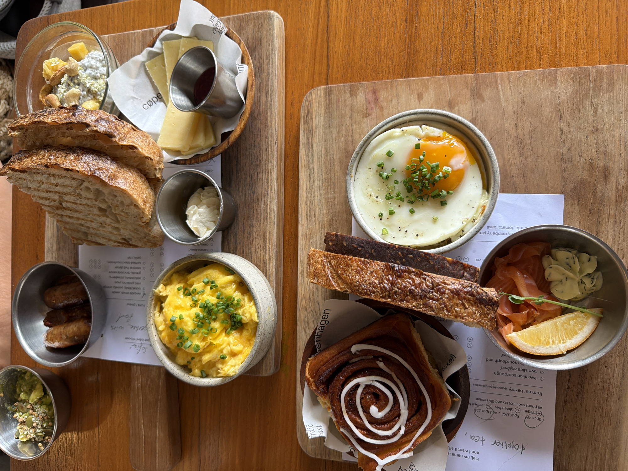 The image shows two wooden serving boards placed on a wooden table. The board on the left contains a glass bowl of muesli, slices of grilled bread, a small wooden basket with sliced cheese and a metal container of jam, a metal container of crème fraîche, a ceramic bowl of scrambled eggs with chives, a metal container of small sausages, and a metal container of an avocado mixture with seeds.
The board on the right contains a metal bowl with a fried egg topped with chives, slices of dark rye bread, a square cinnamon bun with icing, and a metal container with smoked salmon, flavored butter, and a slice of lemon.
The image includes a printed menu sheet placed partially under some of the food items on both boards. Based on the text on the menu, the food is from Copenhagen Bakery in North Kuta, Bali, Indonesia.