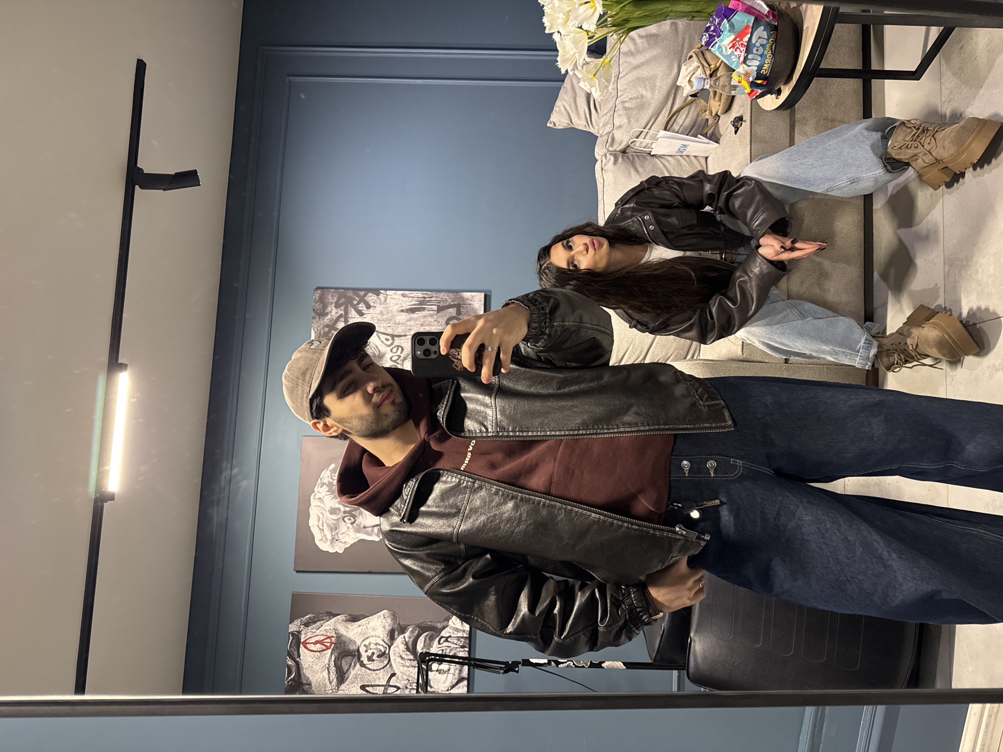 A selfie-style photograph taken in what appears to be an indoor setting. The foreground is dominated by a man wearing a baseball cap, a burgundy hoodie, a black leather jacket, and dark blue jeans. He is holding a smartphone in his right hand, presumably taking the picture. The phone case is dark with what seems to be a leopard print. The background includes a woman reclining on a gray couch, also wearing a black leather jacket and light-colored jeans. Beside the couch is a small black table. The wall is painted a dark blue. Several framed artworks are visible on the wall behind the man. The lighting is a mix of natural and artificial. A tall, thin light fixture is visible near the edge of the mirror. The image gives the impression of a posed, stylish moment captured in an interior space. Yerevan, Armenia is not clearly recognizable from the image.