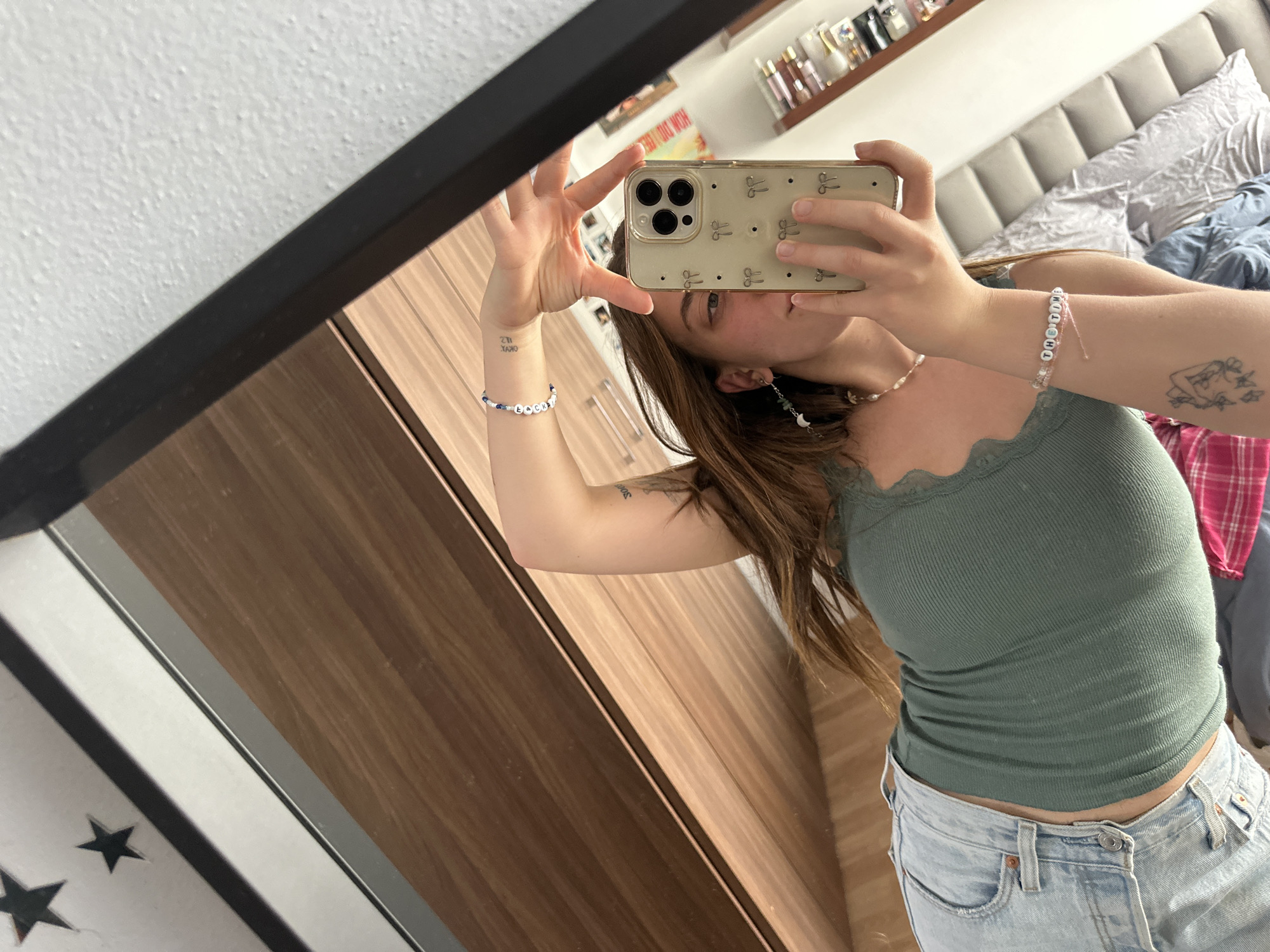 A woman is taking a selfie in front of a mirror. She is holding her phone up with her right hand, obscuring her face. She is wearing a green ribbed tank top and light-wash jeans. Her hair is long and brown. She has bracelets on both wrists and an earring on her left ear. A bed with a grey headboard and bedding is visible in the background. There are shelves with various items on them above the headboard. The mirror has a black frame, and part of a door or wall is visible in the reflection. This image was most likely taken in an interior setting, potentially a bedroom, in Huixquilucan de Degollado, Mexico.