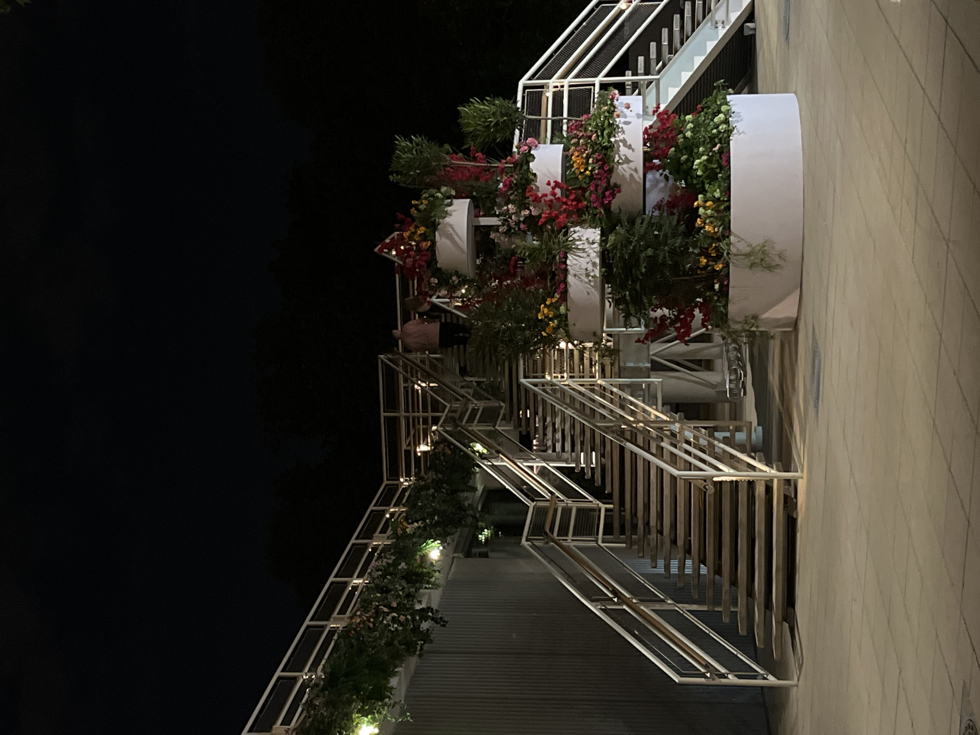 This nighttime image depicts a multi-level outdoor architectural feature adorned with vibrant floral displays. The scene is illuminated by artificial lights, creating a warm glow against the dark sky.
The central elements are modern white and silver metal stairs and railings, which ascend and descend, suggesting multiple accessible levels. Integrated into this structure are large, white, cylindrical and rectangular planters overflowing with lush greenery and colorful flowers. Predominant flower colors include vivid red, pink, and yellow, interspersed with verdant foliage, creating a cascading, layered effect. Small light sources are strategically placed within the planting arrangements, highlighting the flora.
On the lower left, partially obscured by the foliage, a person wearing a light-colored top and dark pants can be seen near the stairs, suggesting movement or presence within the space. To the far right, a light-colored, tiled paved surface is visible.
The overall impression is that of an inviting and beautifully decorated public or semi-public space, possibly a terraced garden, promenade, or a part of a building complex designed for evening ambiance. The time of day is clearly night, with a dark, unlit background on the left. There is no visible text in the image. The location context for this scene is Moscow, Russia.