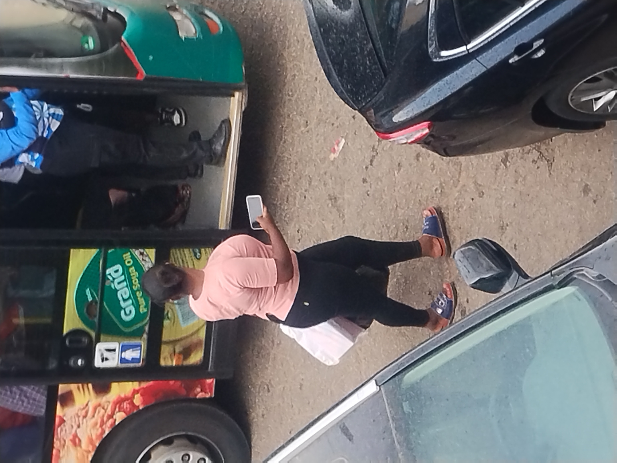 The image shows a street scene with several vehicles and pedestrians. A woman wearing a pink shirt and black leggings is standing and looking at her mobile phone. She is next to a vehicle displaying "Grand Pure Soya Oil" advertisement. Another pedestrian is seated inside the vehicle. Two cars are parked nearby. The location is in an urban environment.