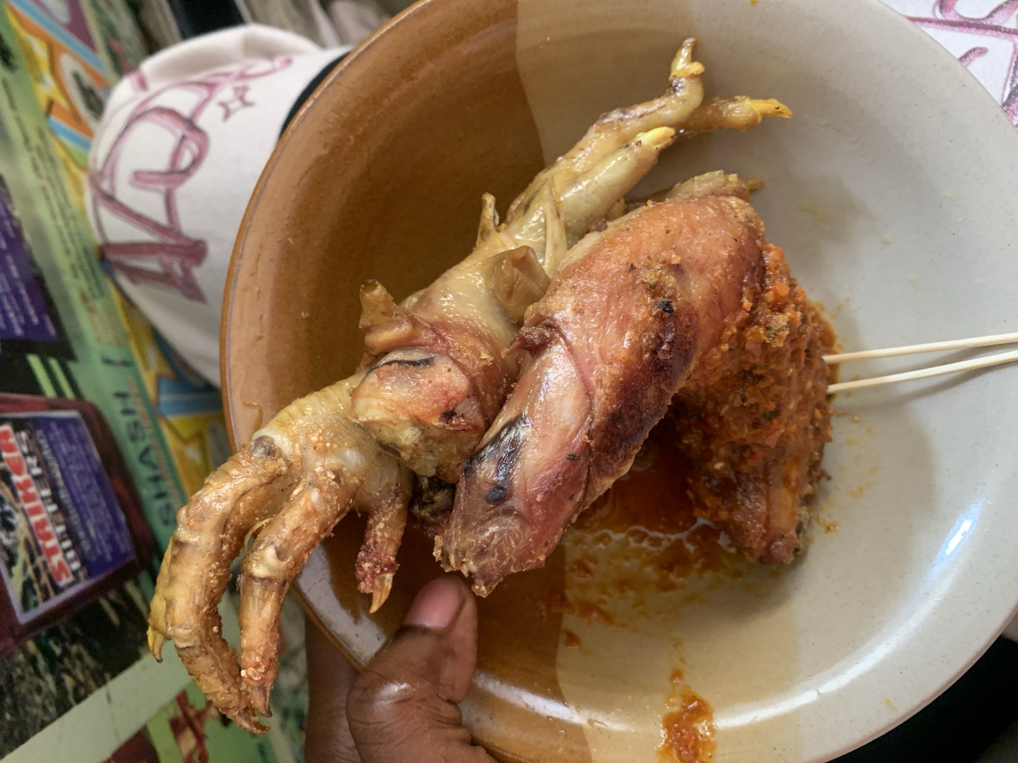 The image shows a close-up of a brown and beige ceramic bowl held by a dark-skinned hand. The bowl contains three pieces of what appears to be cooked chicken, two of which are chicken feet. One piece of chicken is on a wooden skewer. There is a small amount of sauce in the bottom of the bowl. The bowl sits on a colorful surface, possibly a table or tablecloth, with a white object with pink markings partially visible in the background. The location is possibly Jos, Nigeria.