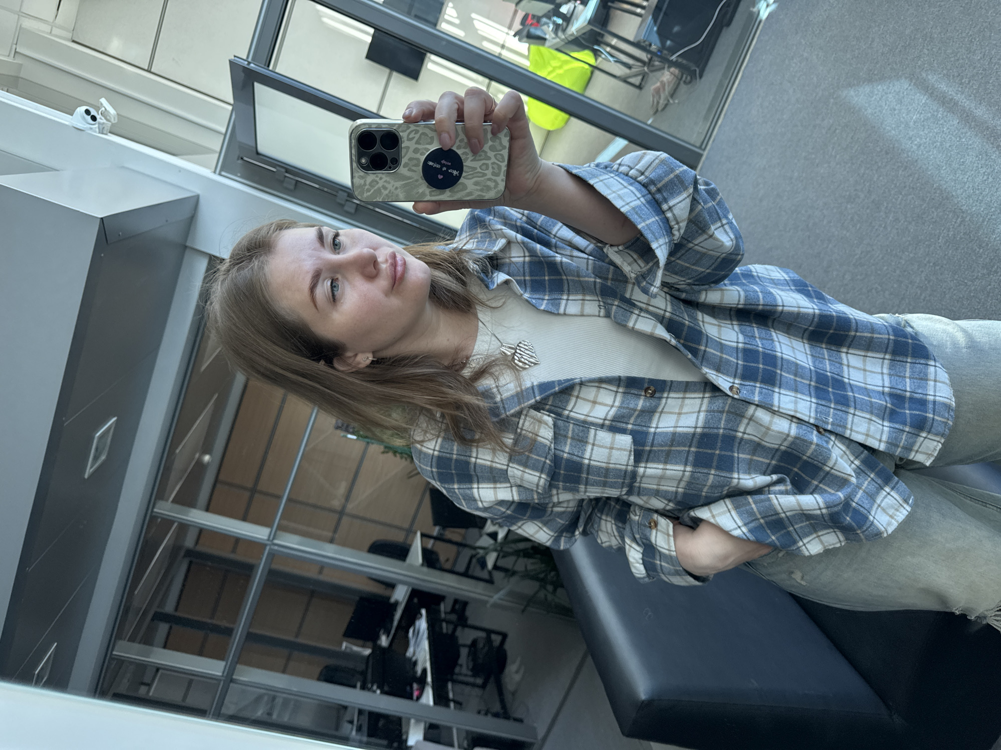 A young woman takes a selfie in a room with large windows. She is wearing a blue and white plaid shirt over a white top and light-colored jeans. She holds a smartphone with a patterned case. Outside the windows, there are office spaces visible. The location is Perm, Russia.