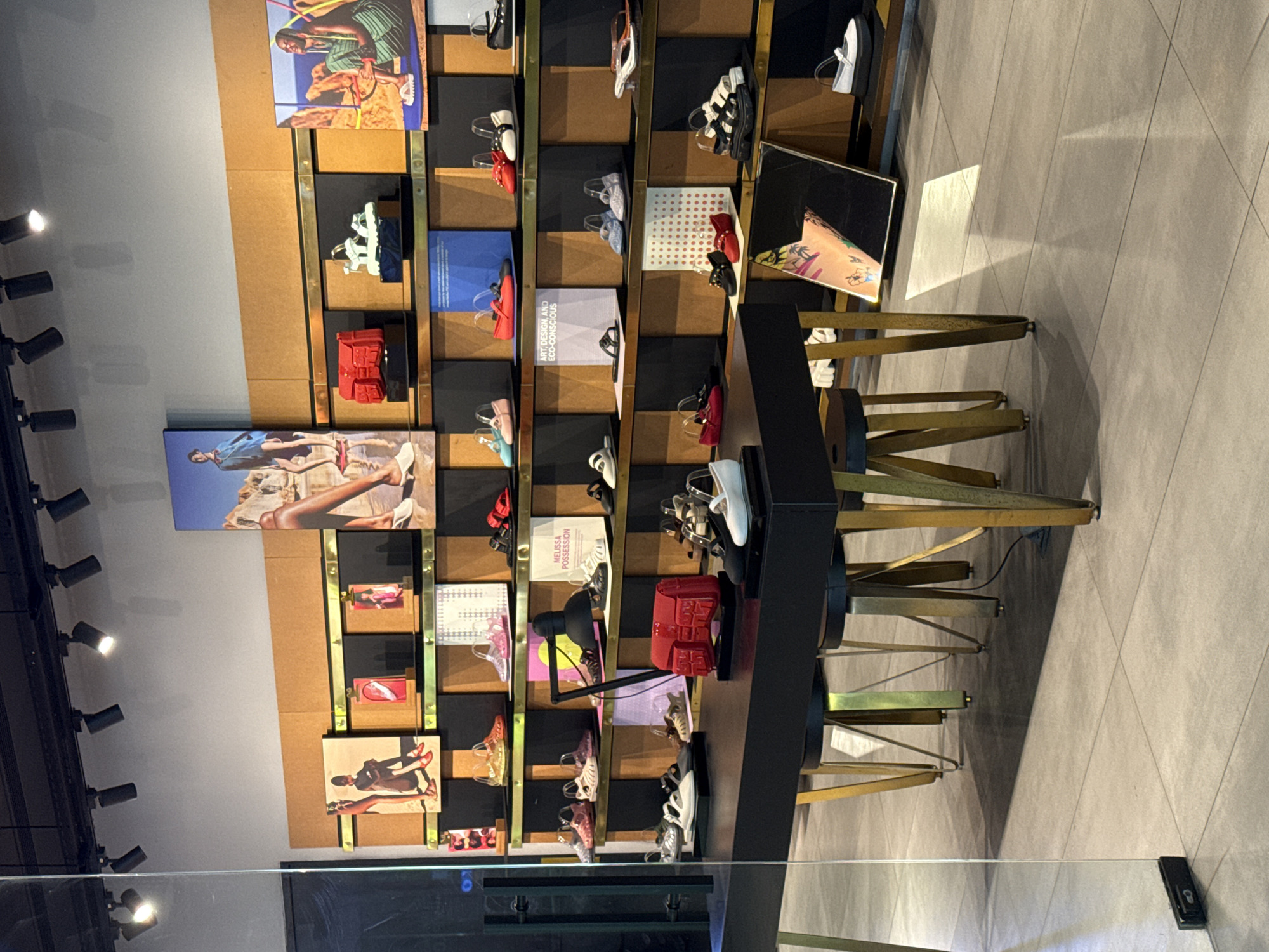 The image shows a retail display for footwear, possibly sandals and shoes, in what appears to be a store in Kuta, Indonesia. The display unit is multi-tiered with various shelves showcasing the footwear alongside small artworks or promotional materials. A black desk-like structure stands in front of the display, supported by gold-colored geometric legs. A desk lamp provides lighting on the merchandise. The scene is viewed through what is likely a glass barrier or display window, creating a slight reflection. The store's interior is modern and well-lit, with track lighting visible on the ceiling. The flooring is composed of large grey tiles. The picture appears to be taken from a slightly elevated position.